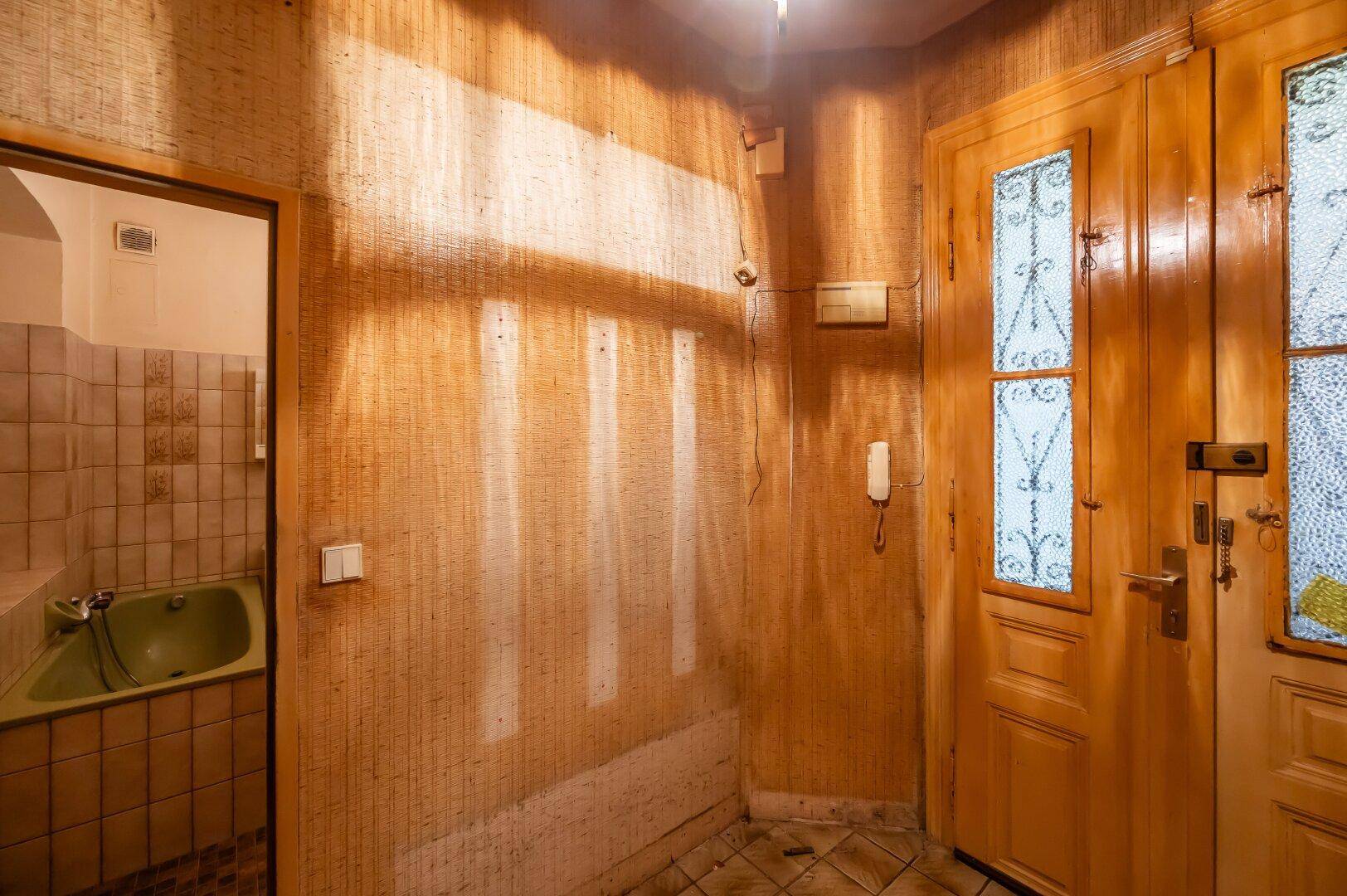 Flur mit gemusterter Tapete und einer Holztür, die zu einem Badezimmer mit grüner Wanne führt.