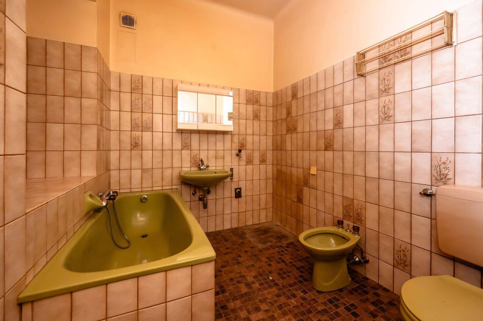 Badezimmer im Stil der 80er Jahre mit grüner Badewanne, Toilette und Waschbecken sowie beigefarbenen Fliesen.