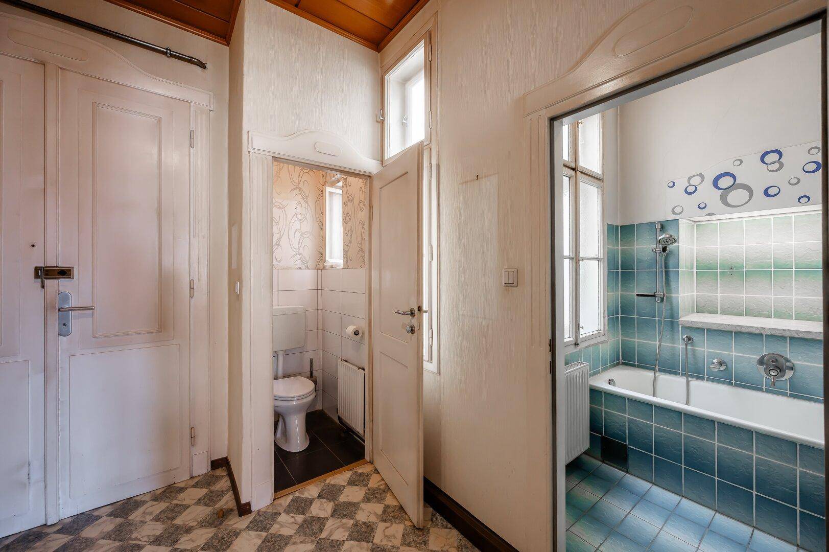 Flur mit Schachbrettmuster-Fliesenboden, Zugang zu einem WC und einem Badezimmer mit Badewanne.
