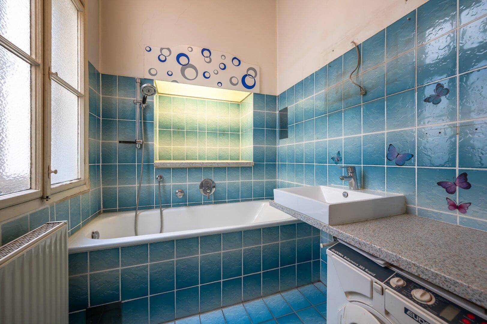 Badezimmer mit blauen Fliesen, Badewanne, Waschbecken und Waschmaschine unter der Arbeitsplatte.