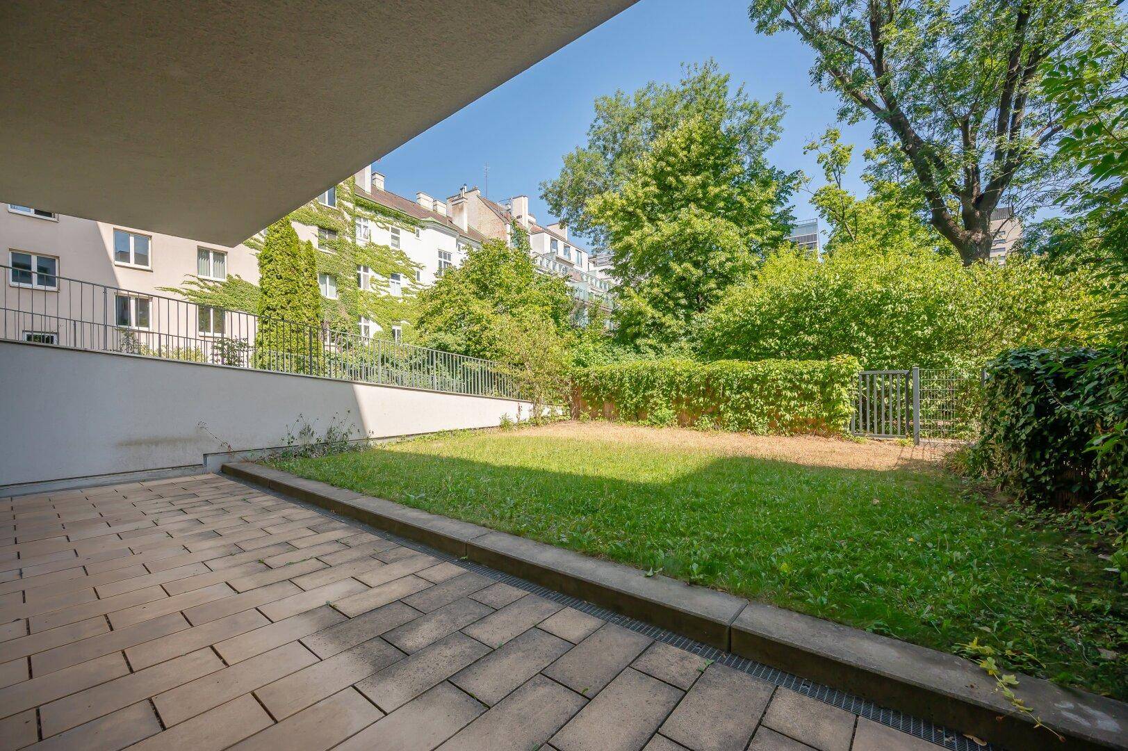 Sonniger Gartenbereich mit gepflasterter Terrasse und üppigem Grün in urbaner Umgebung.