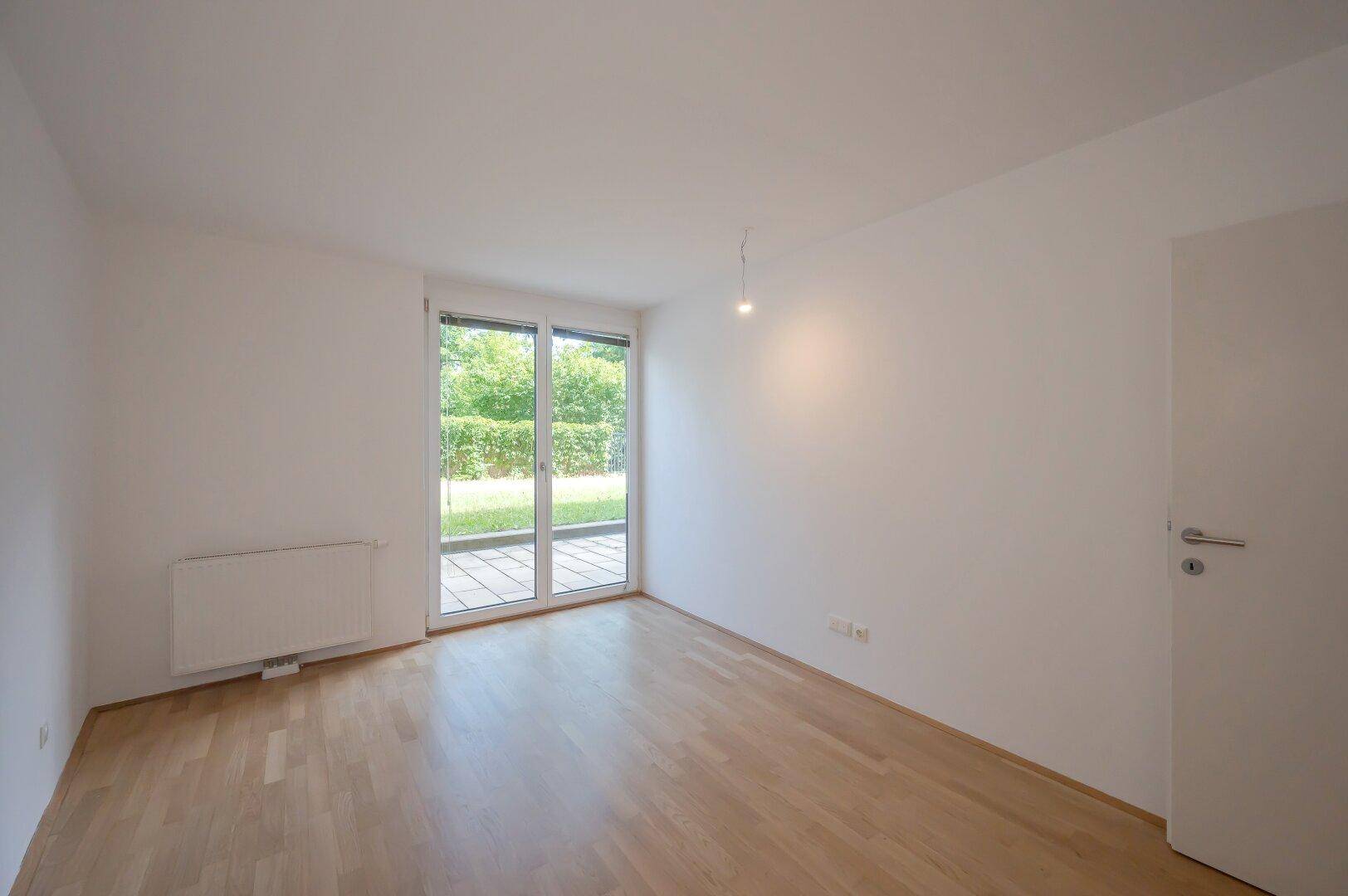 Lichtdurchfluteter Raum mit Holzboden und großem Fenster mit Blick ins Grüne.