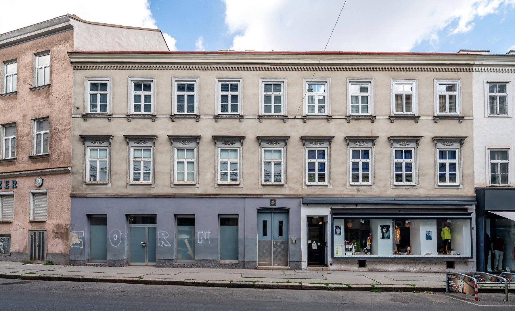 Fassade eines älteren Gebäudes mit vielen Fenstern und Ladenlokalen im Erdgeschoss an einer Stadtstraße.