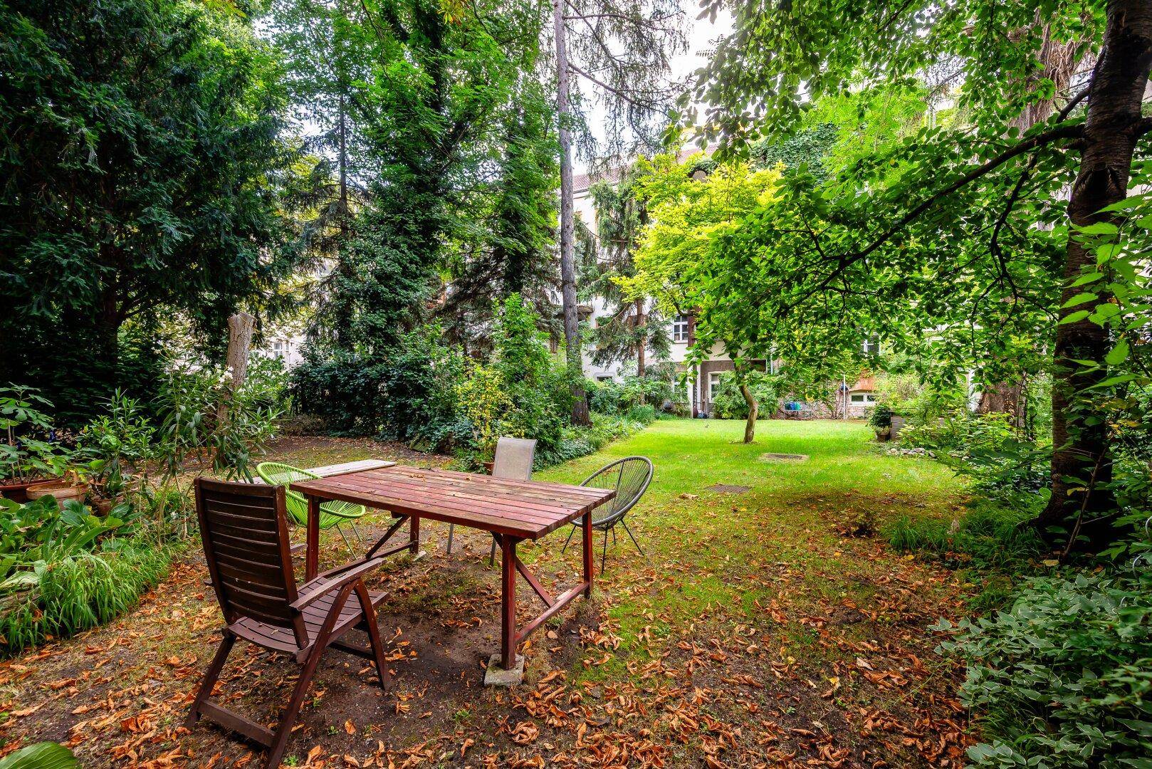 Ruhiger Gartenbereich mit Holztisch und Stühlen unter schattenspendenden Bäumen.