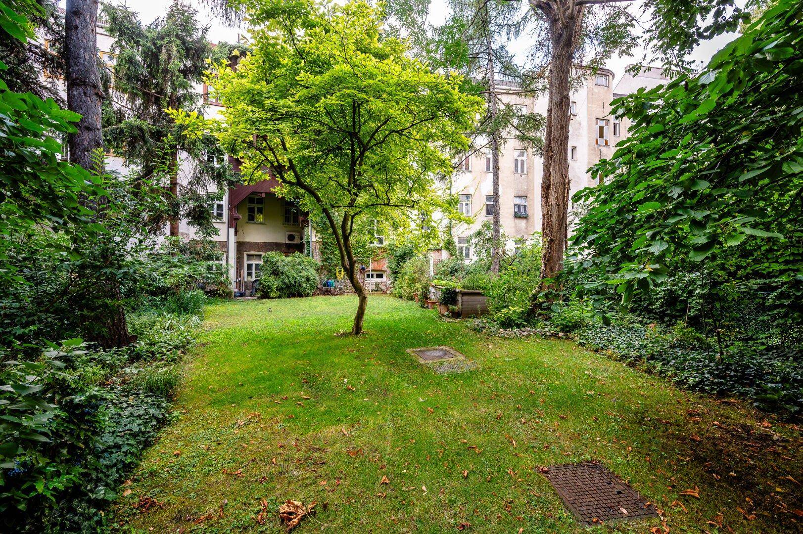 Weitläufiger Garten mit Rasenfläche und altem Baumbestand, umgeben von Gebäuden.