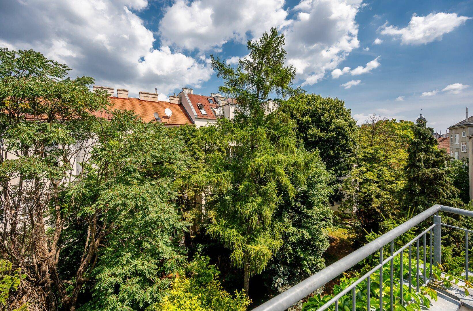 Blick von einem Balkon über grüne Bäume auf die Dächer umliegender Gebäude unter blauem Himmel.