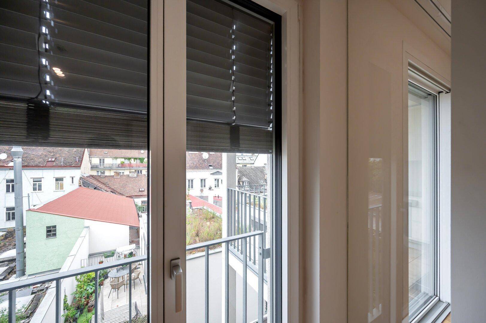 Fenster mit Jalousien und Blick auf die umliegenden Dächer und Balkone.