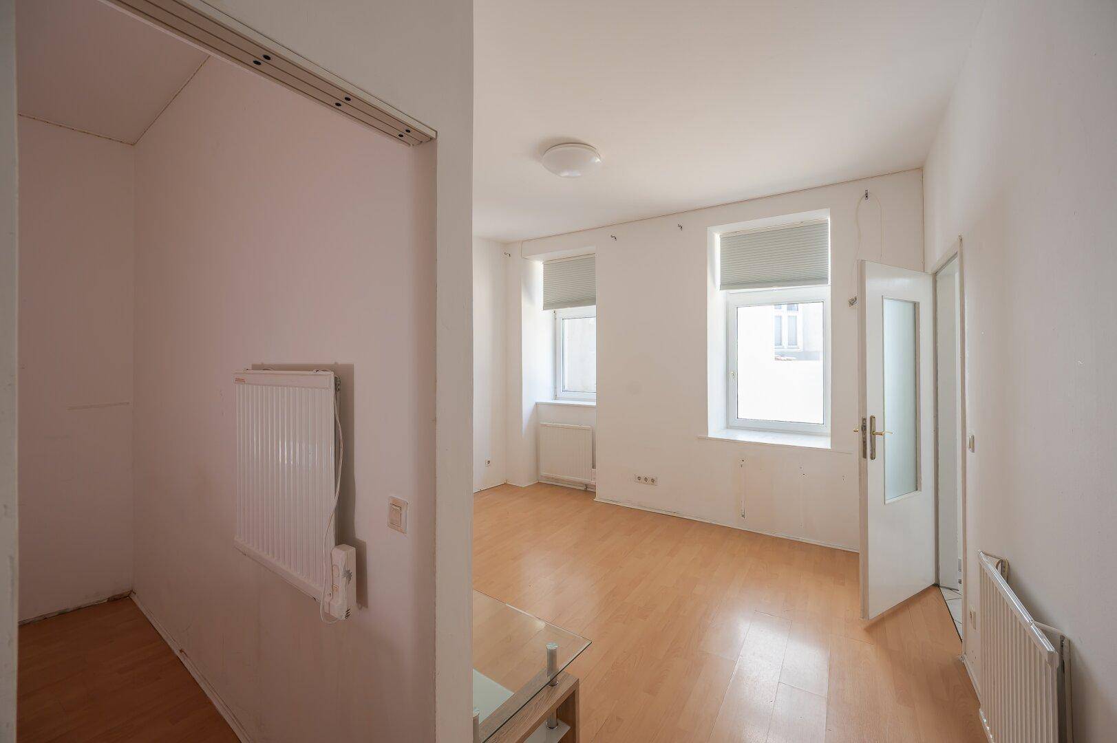 Blick vom Flur in ein helles Zimmer mit Parkettboden, weißen Wänden und zwei Fenstern mit Jalousien.