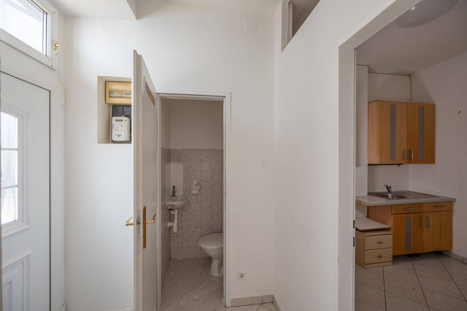Eingangsbereich mit Fliesenboden, Zugang zu einer Toilette und Blick auf eine kleine Küchenzeile.