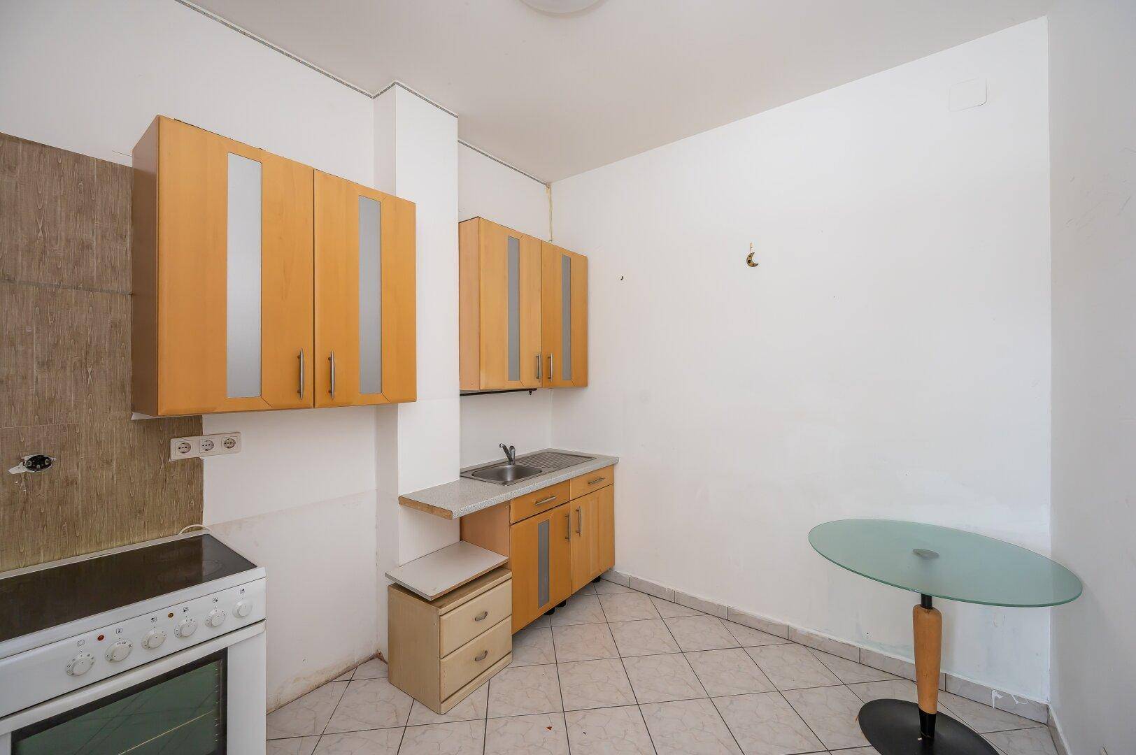 Einfache Küchenzeile mit Holzschränken, Spüle, Herd und Fliesenboden, ideal für kleine Wohnungen.