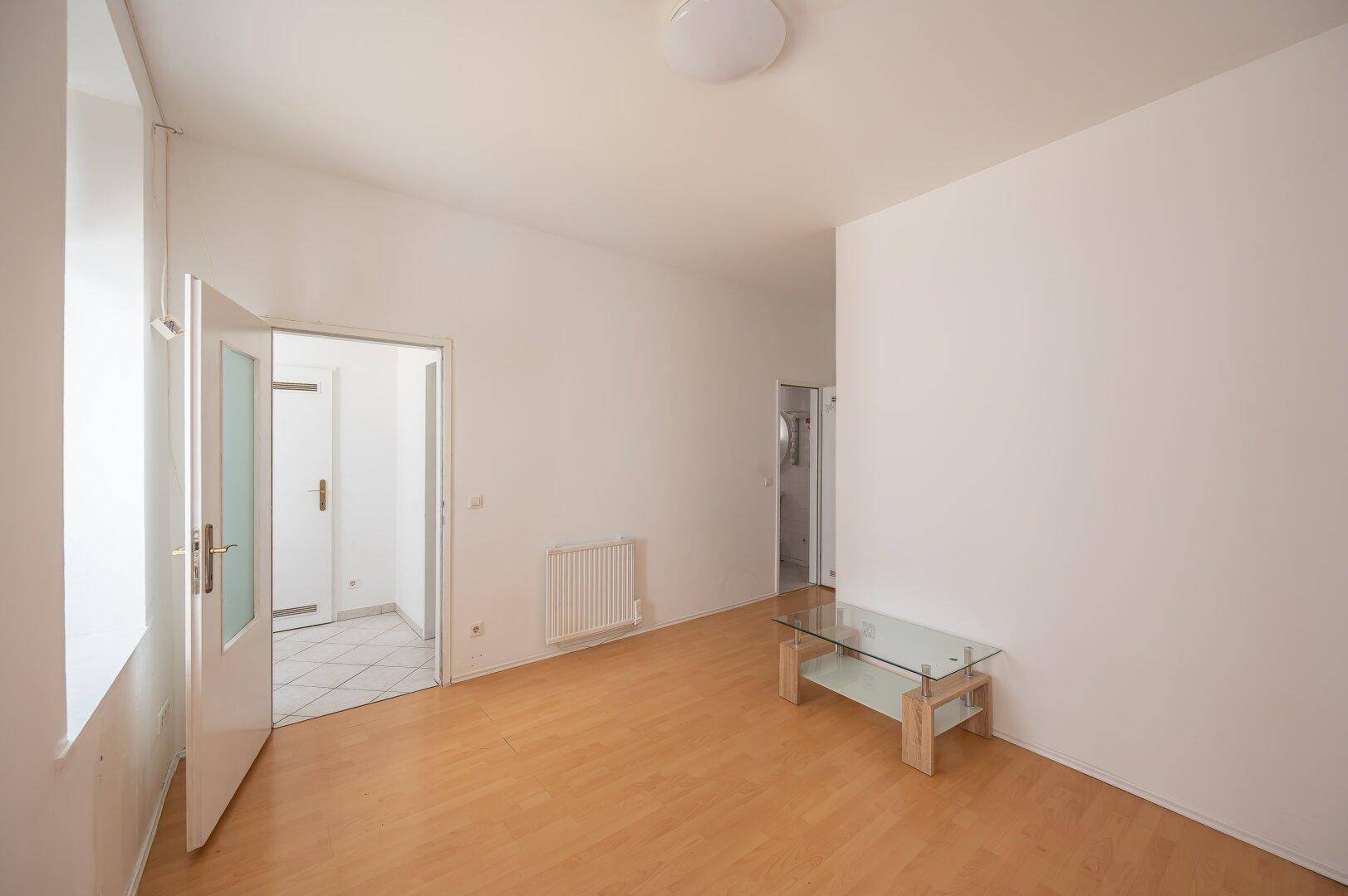 Helles Wohnzimmer mit Parkettboden und Glastisch, mit Zugang zu einem gefliesten Bereich.