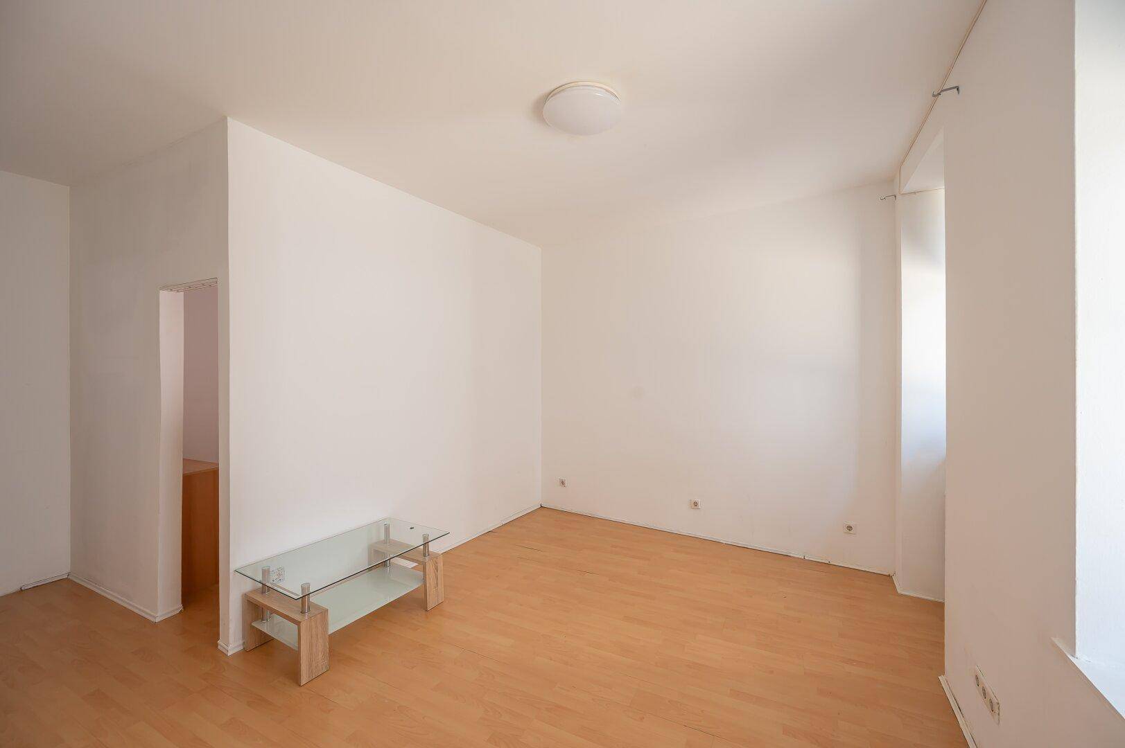 Geräumiges Wohnzimmer mit hellem Parkettboden und minimalistischem Glastisch, offen zu einem weiteren Bereich.
