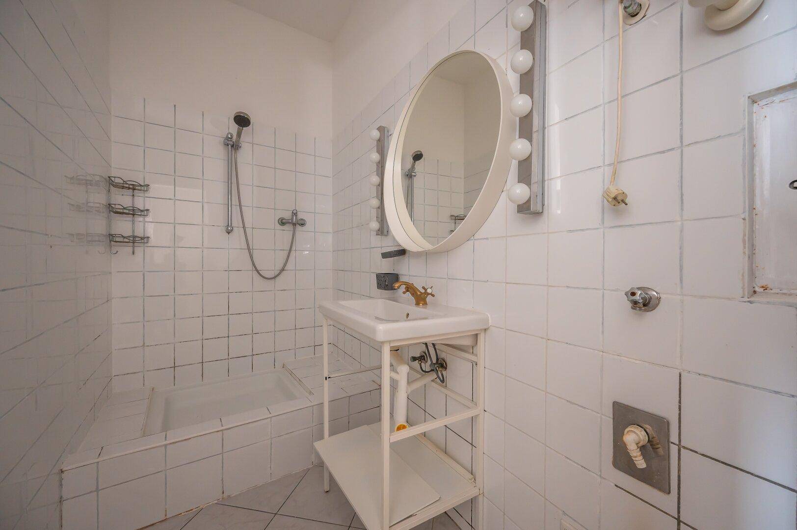 Gefliestes Badezimmer mit Dusche, Waschbecken und ovalem Spiegel mit Beleuchtung, in schlichtem Design.