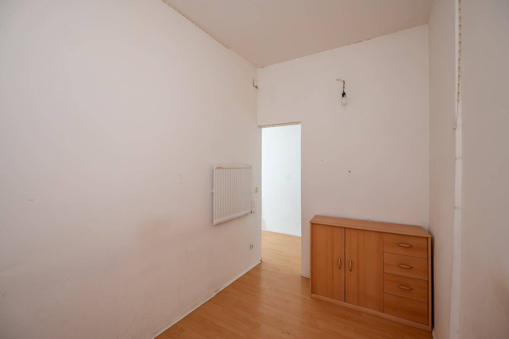 Leeres Zimmer mit hellem Parkettboden und weiß gestrichenen Wänden, mit einem kleinen Holzschrank.