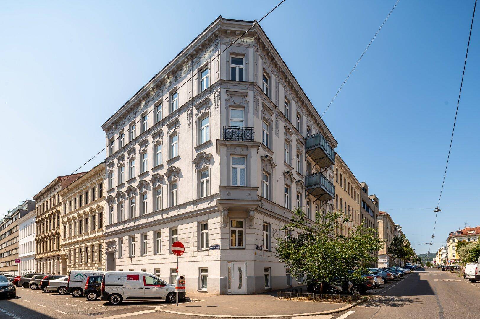 Historisches Eckgebäude mit Balkonen an einer belebten Stadtstraße unter blauem Himmel.