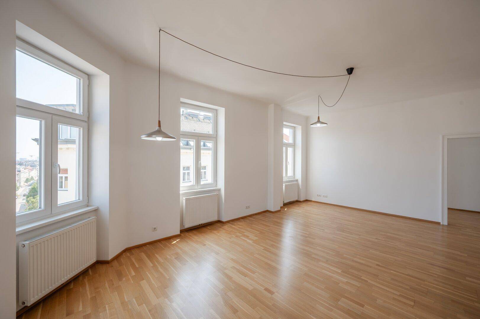 Geräumiger, heller Wohnbereich mit Parkettboden und großen Fenstern, die viel Tageslicht hereinlassen.