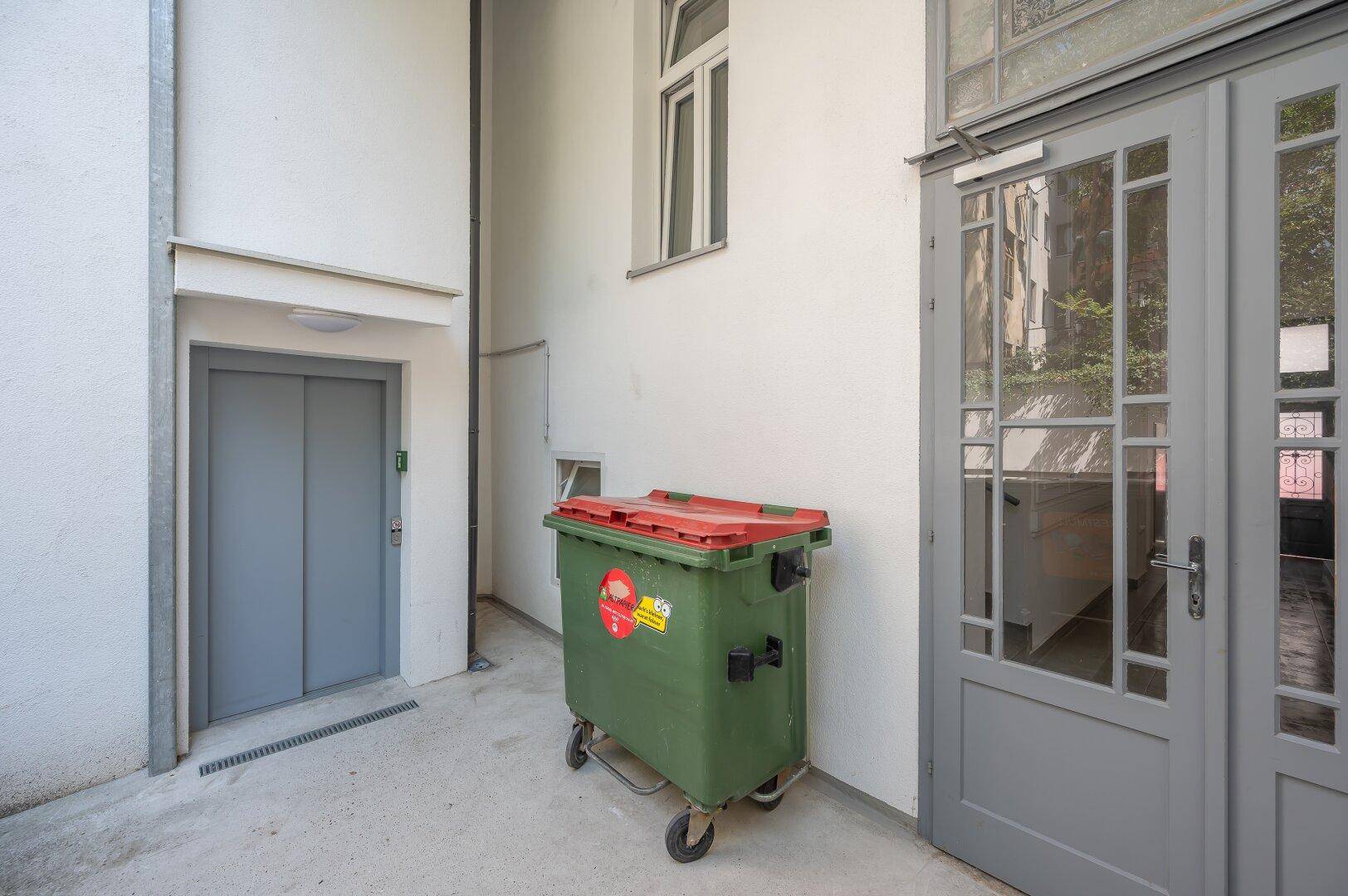Außenbereich eines Gebäudes mit Aufzugzugang und einem großen Müllcontainer.