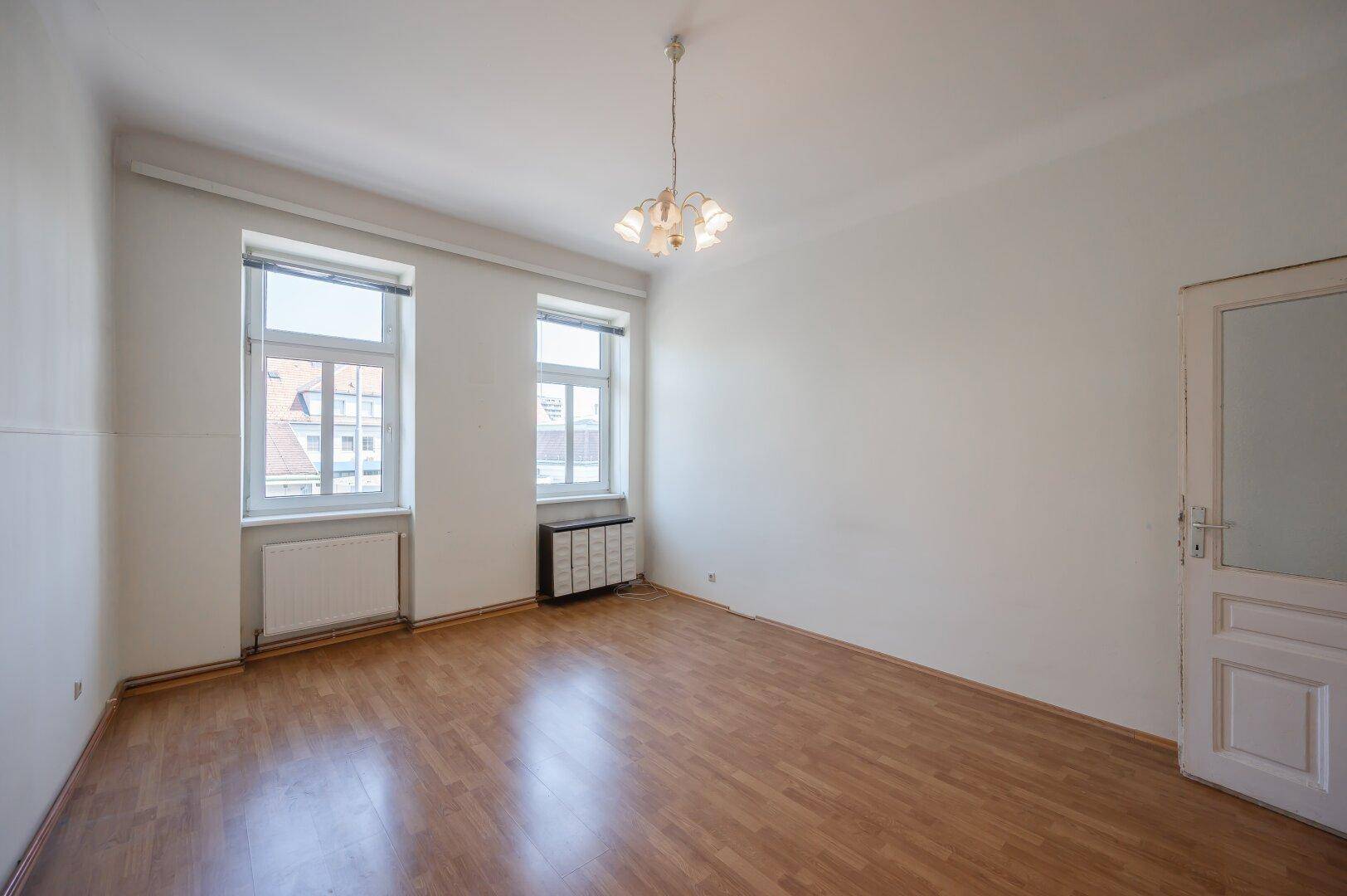 Leeres Zimmer mit Laminatboden, zwei Fenstern und einfacher Deckenleuchte.