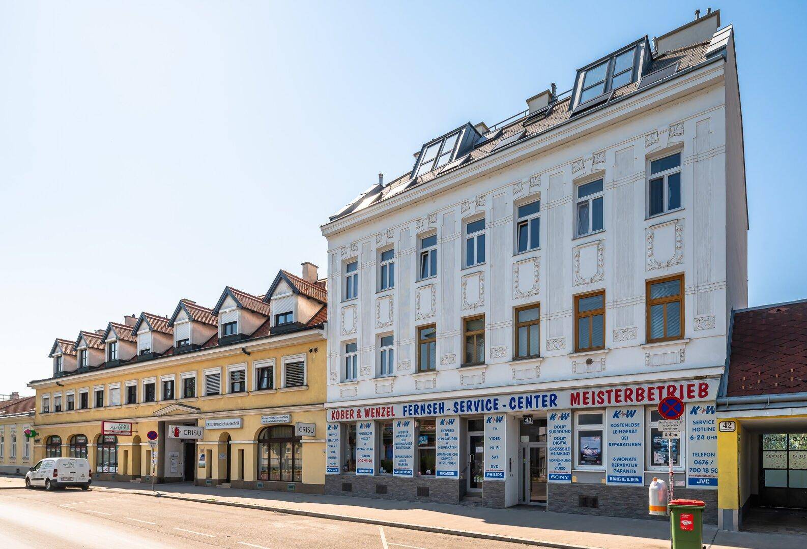 Straßenansicht mit mehreren Gebäuden und Geschäftsfassaden unter blauem Himmel.