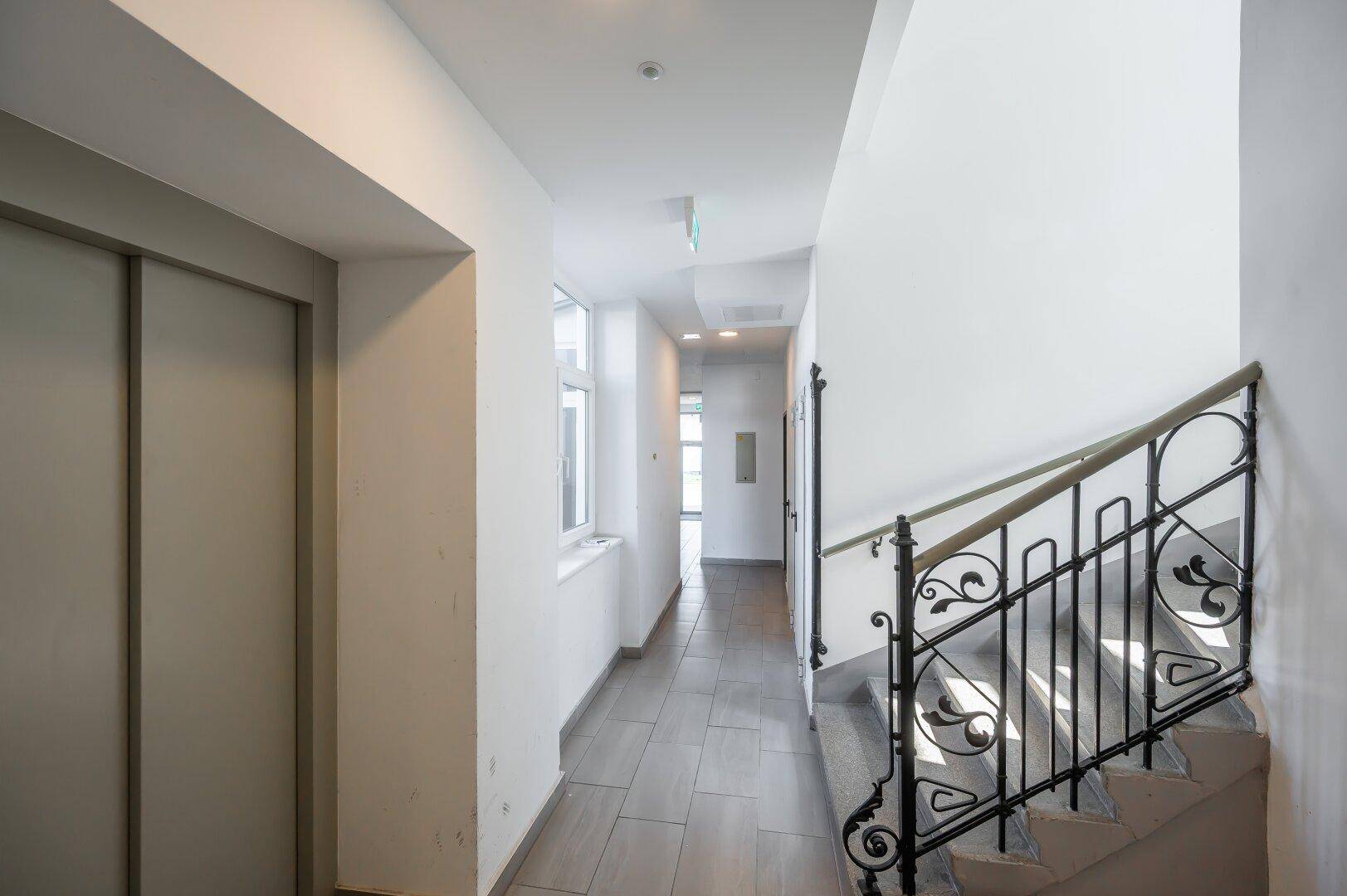 Gemeinschaftsflur mit Fliesenboden, Treppe mit schmiedeeisernem Geländer und Aufzug.