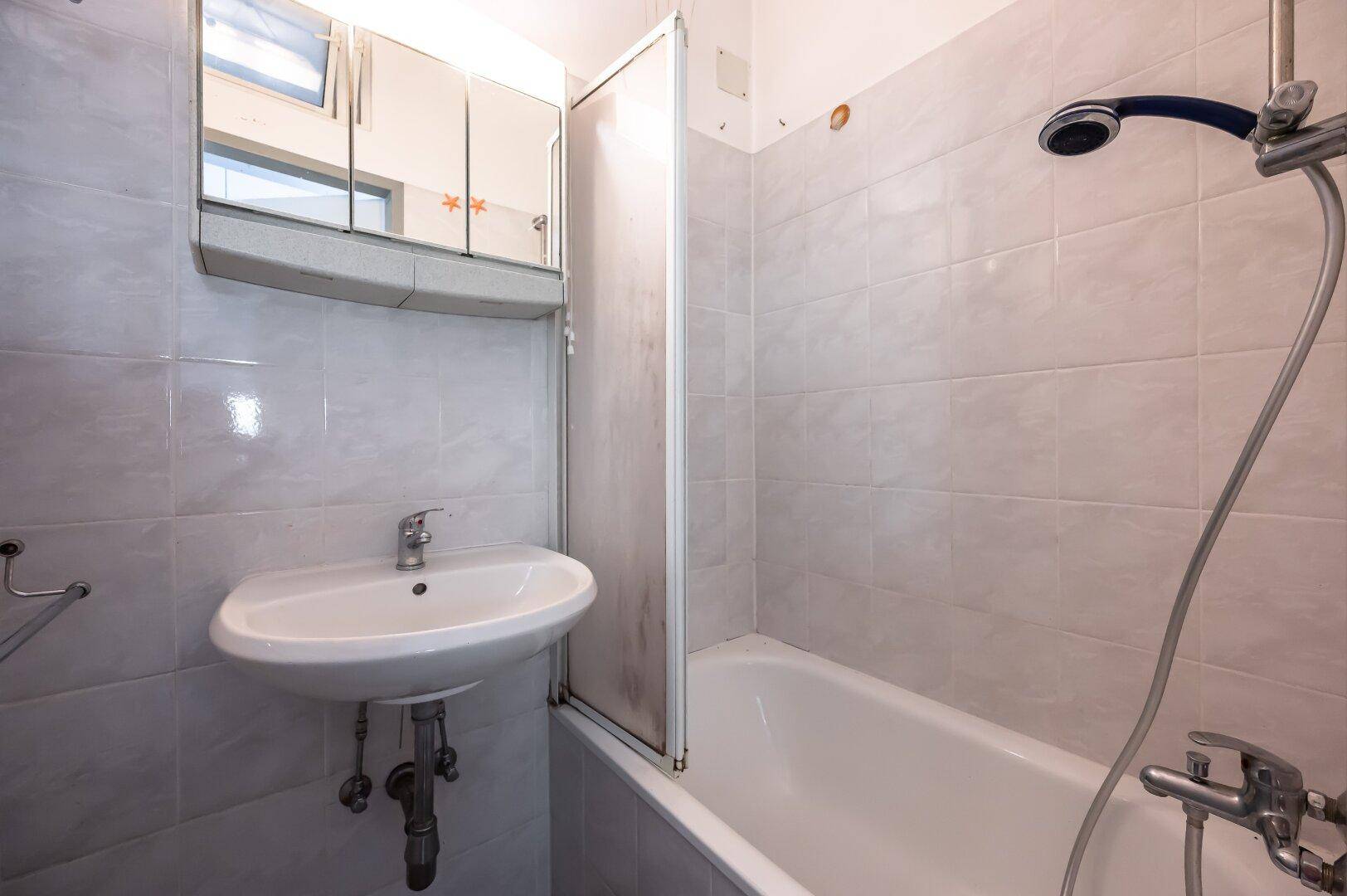 Badezimmer mit hellgrauen Fliesen, Badewanne mit Duschfunktion und Waschbecken.