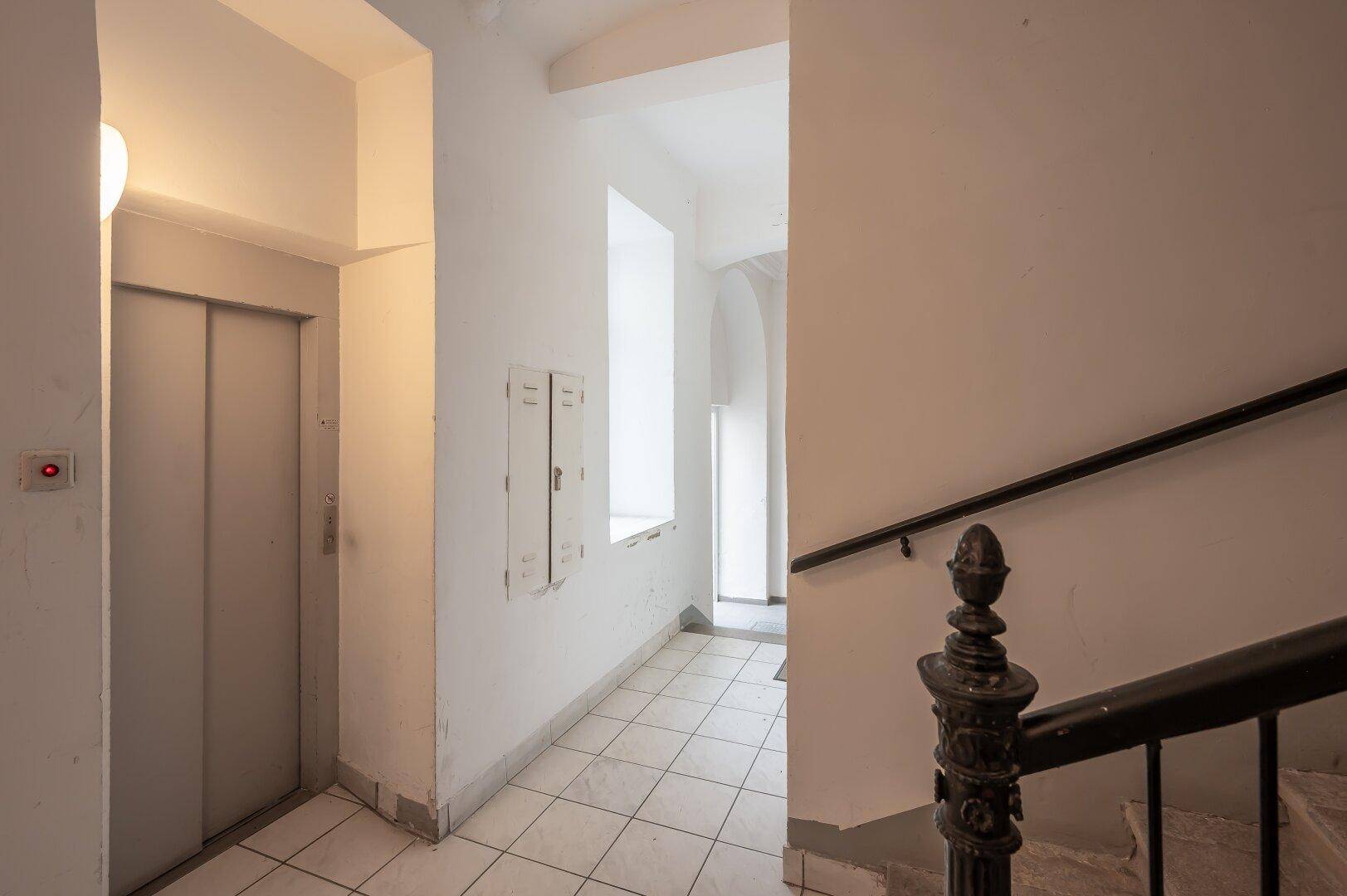 Gemeinschaftlicher Flur mit Aufzug und Treppenhaus in einem Altbau mit Fliesenboden.