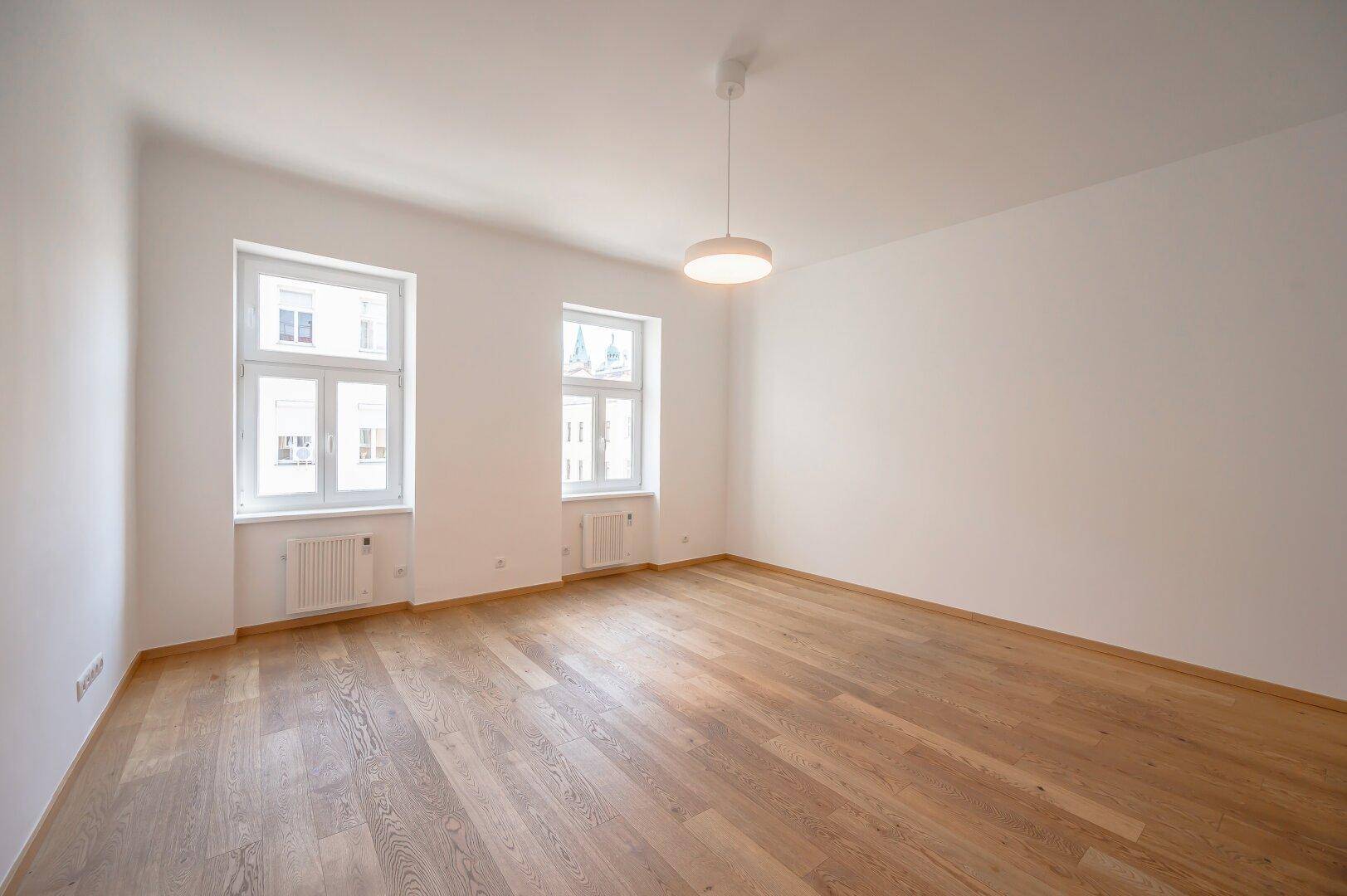 Leeres, helles Zimmer mit Holzboden und zwei großen Fenstern.