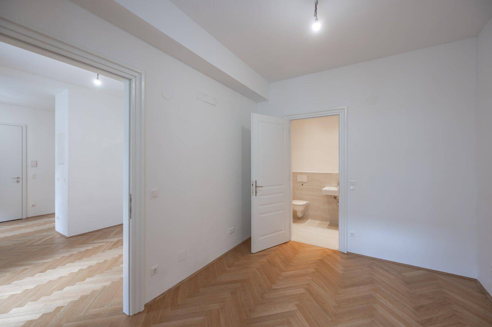 Helles Zimmer mit Fischgrätparkett und Zugang zu einem separaten WC.