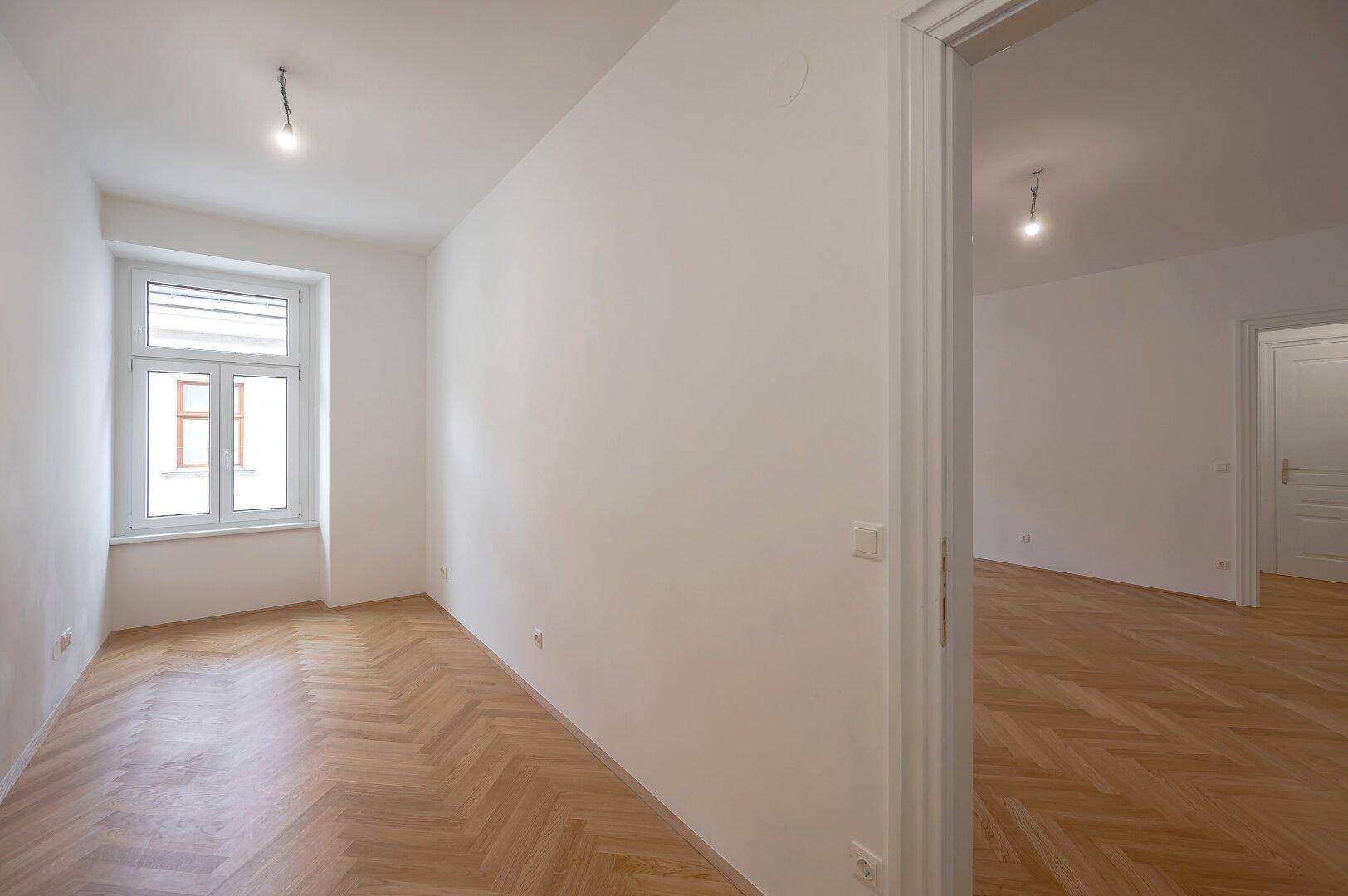 Geräumiges Zimmer mit Fischgrätparkett und Fenster, das viel Tageslicht hereinlässt.