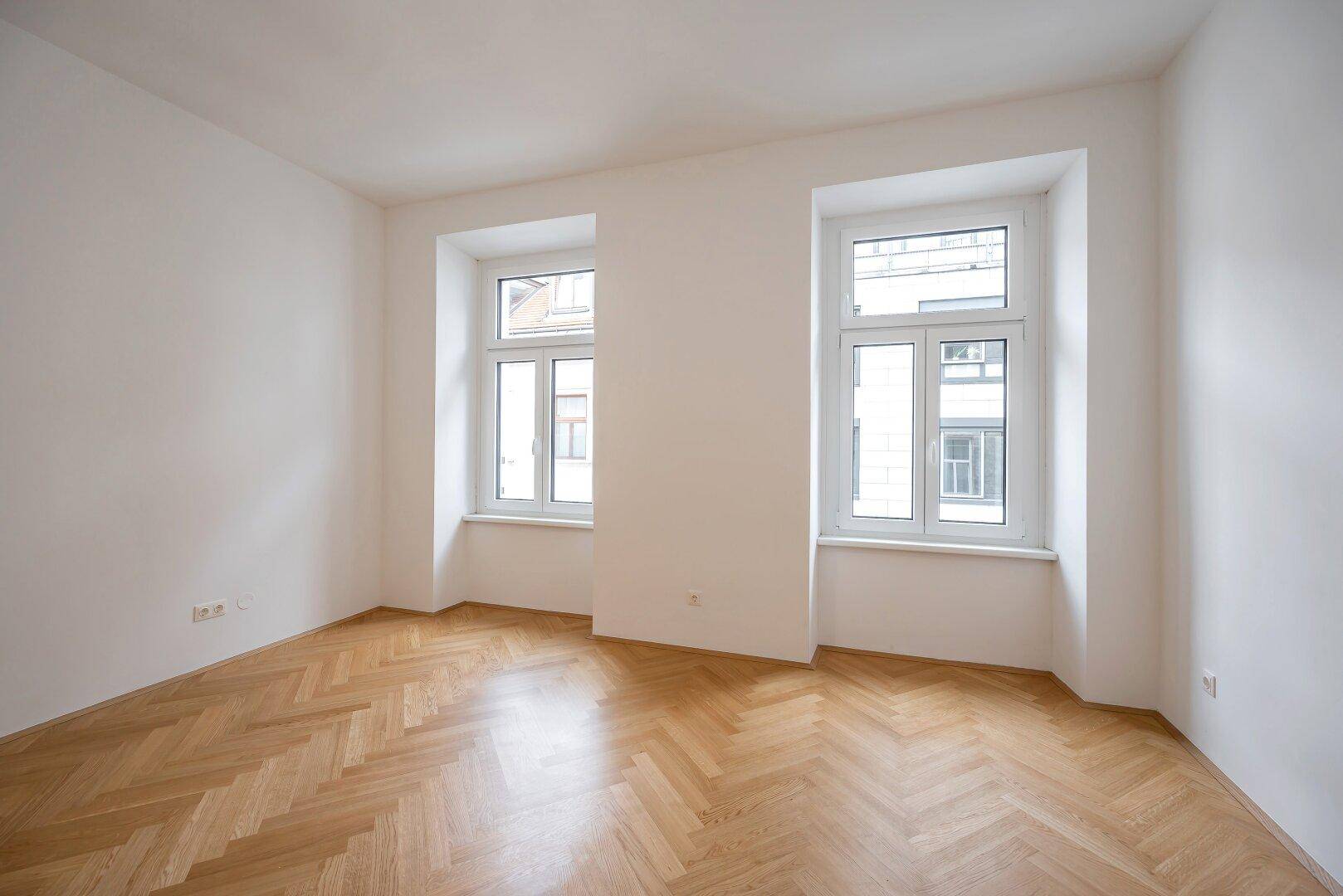 Helles und leeres Zimmer mit zwei großen Fenstern und klassischem Fischgrätparkett.