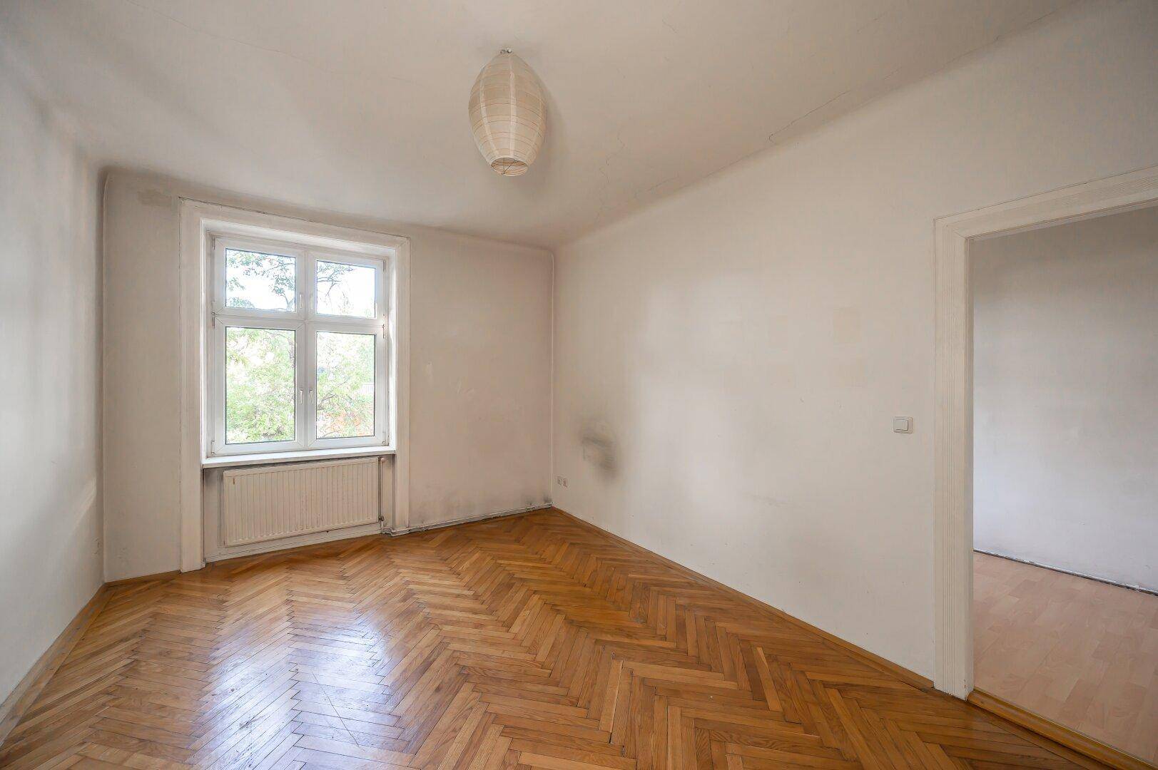 Leeres Zimmer mit Fischgrätparkett und einem Fenster, das viel Tageslicht hereinlässt.