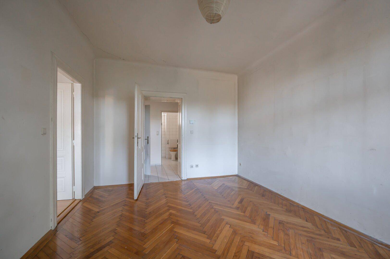 Leeres Zimmer mit Fischgrätparkett und Zugang zu einem weiteren Raum.