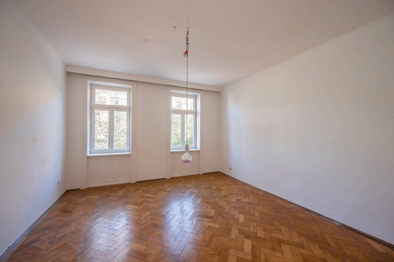 Heller, leerer Raum mit Fischgrätparkettboden und zwei großen Fenstern.