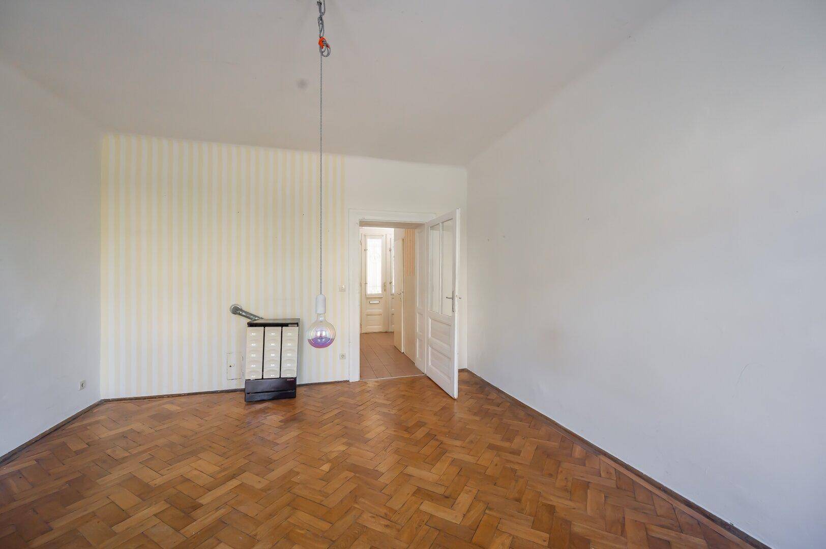 Geräumiges Zimmer mit Fischgrätparkett, einem Kachelofen und Zugang zu weiteren Räumen.