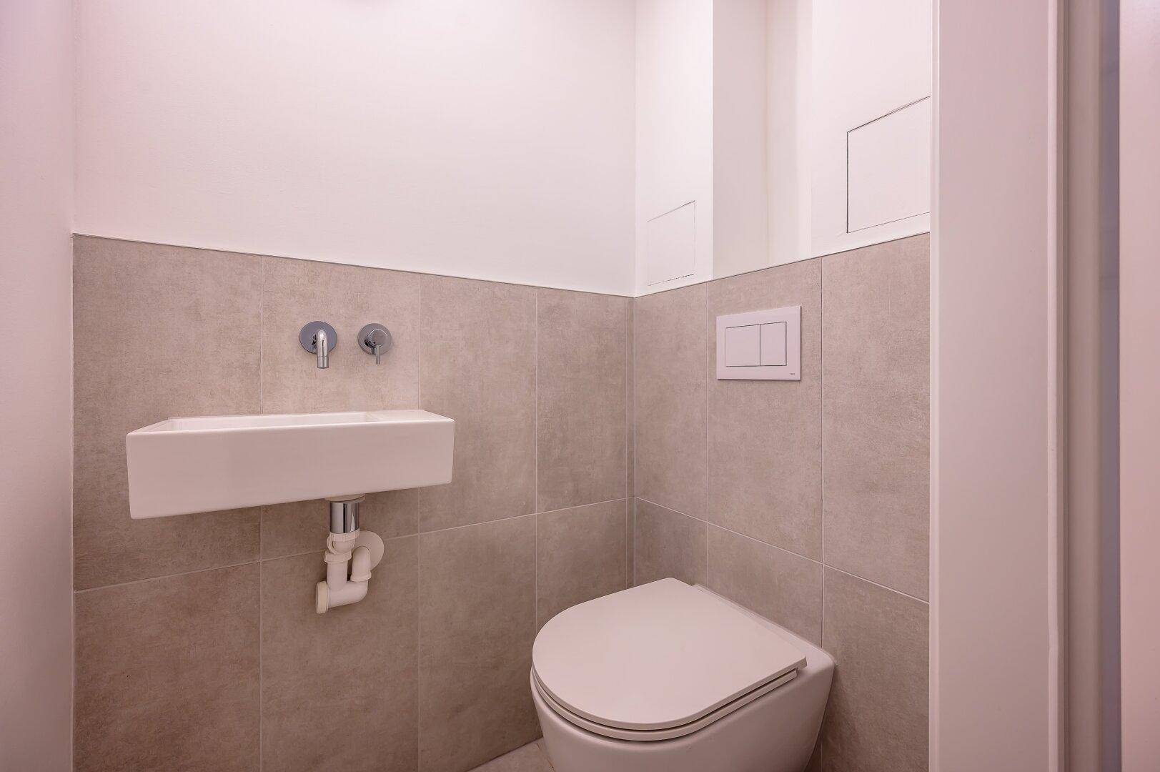 Modernes Gäste-WC mit Wand-WC und kleinem Waschbecken, hell gefliest und funktional.