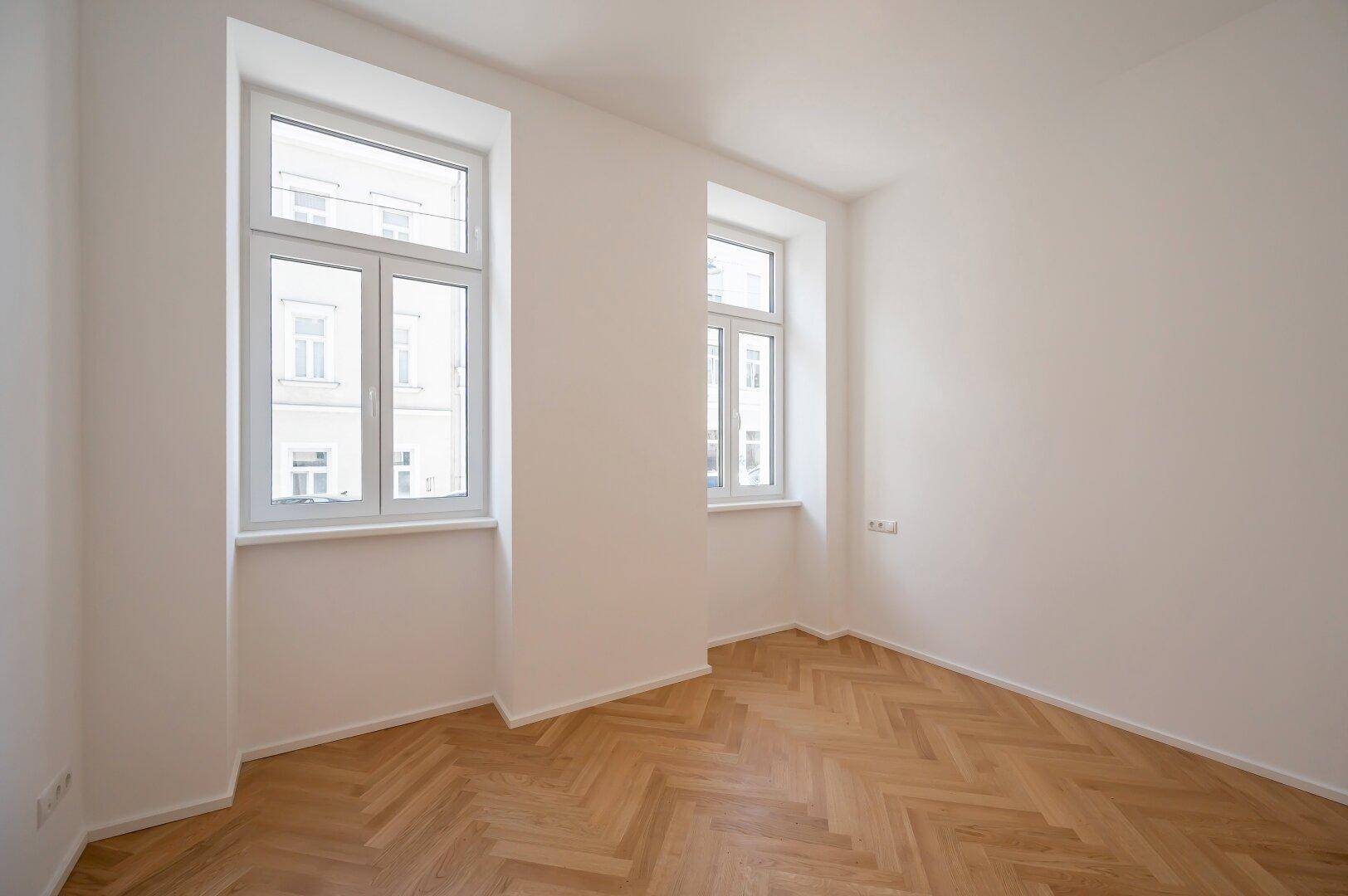 Helles Zimmer mit zwei Fenstern und klassischem Fischgrätparkett, ideal als Schlafzimmer oder Büro.