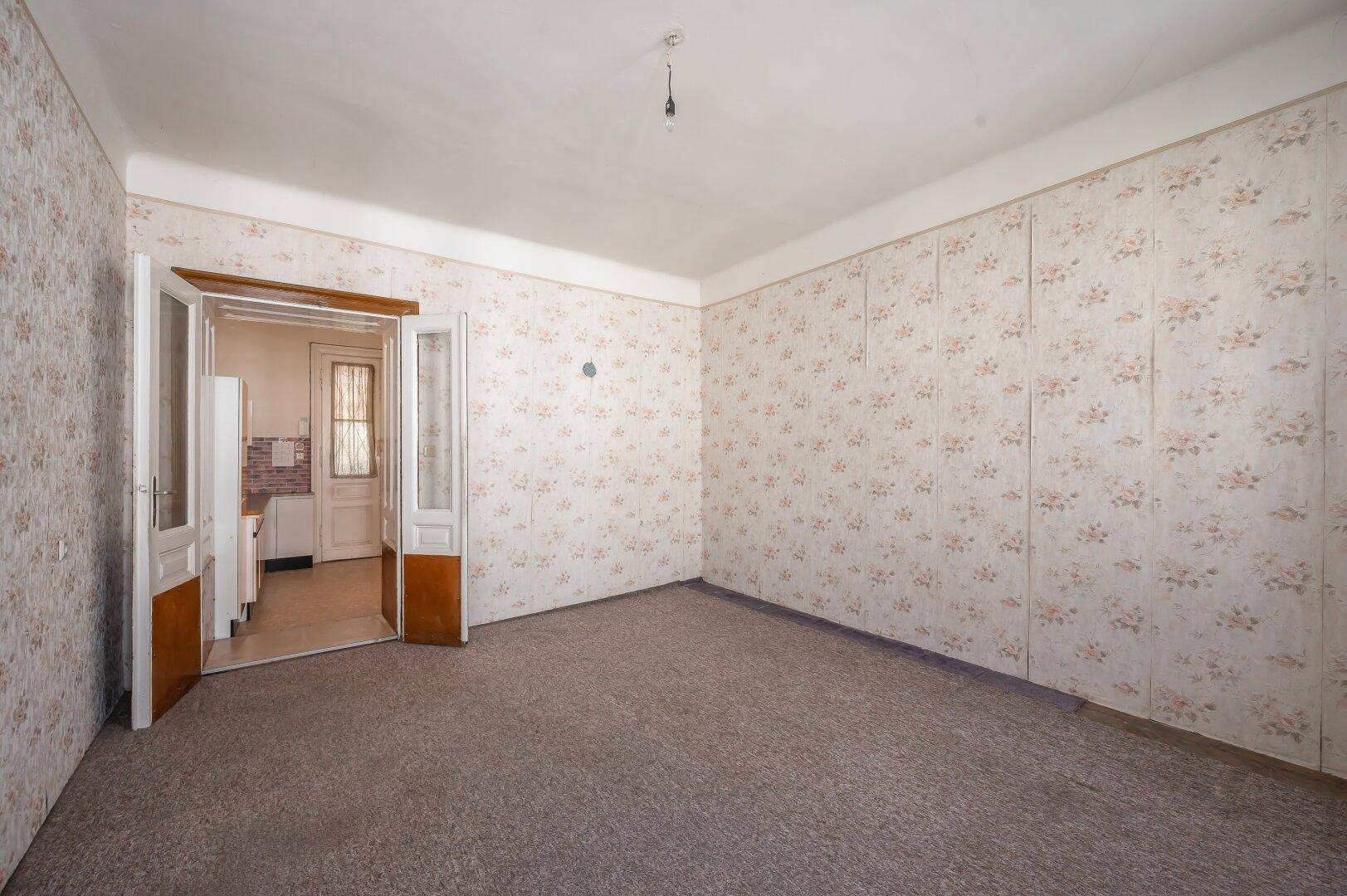 Helles Zimmer mit floraler Tapete und Teppichboden, mit offenem Zugang zur Küche.