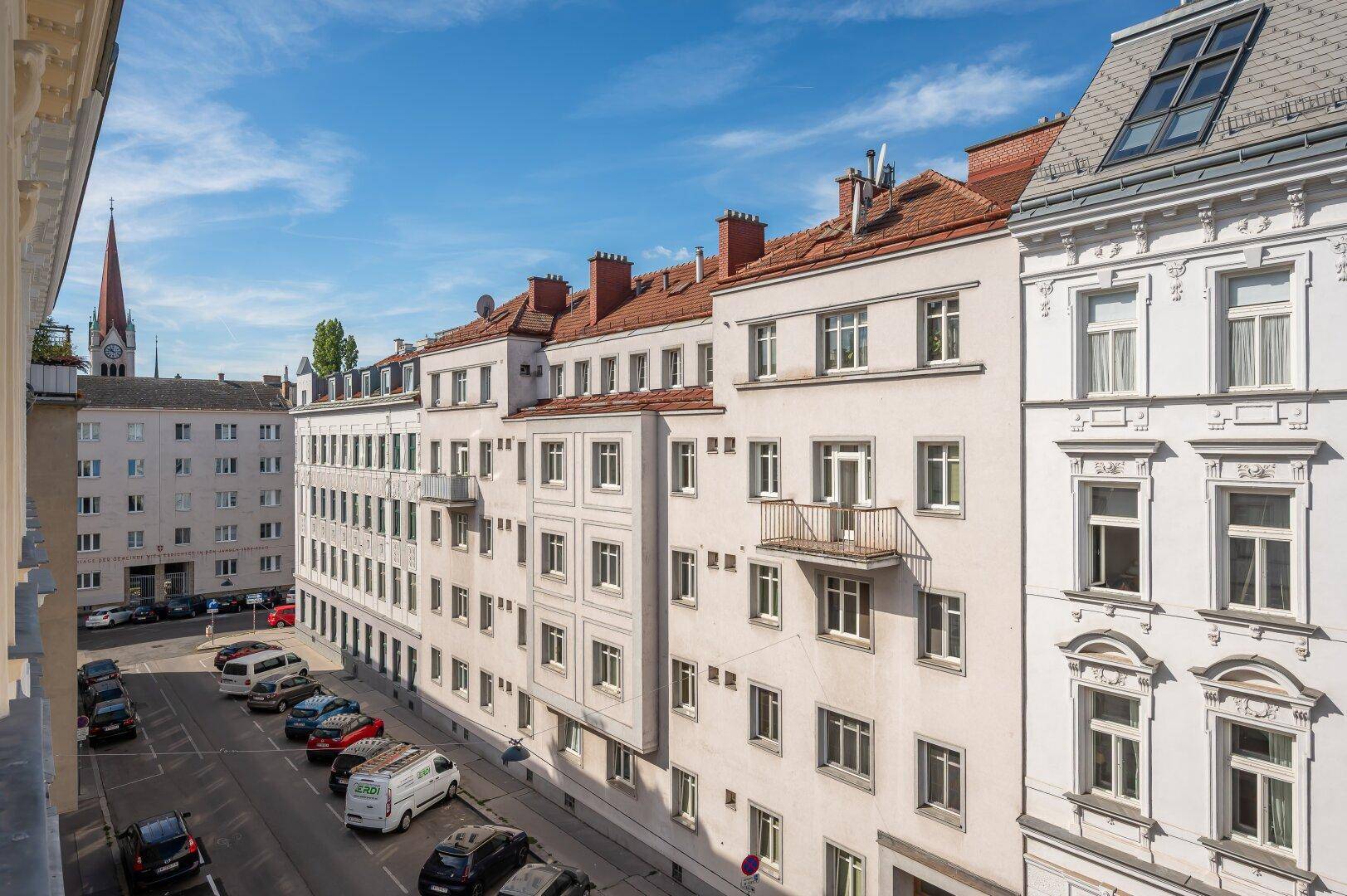 Panoramablick auf eine belebte Straße mit Wohngebäuden und einem Kirchturm in der Ferne.