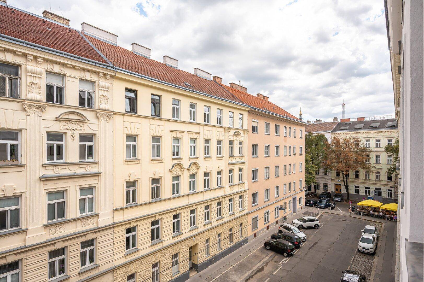 Ansicht einer Straße mit historischen Gebäuden und parkenden Fahrzeugen unter bewölktem Himmel.