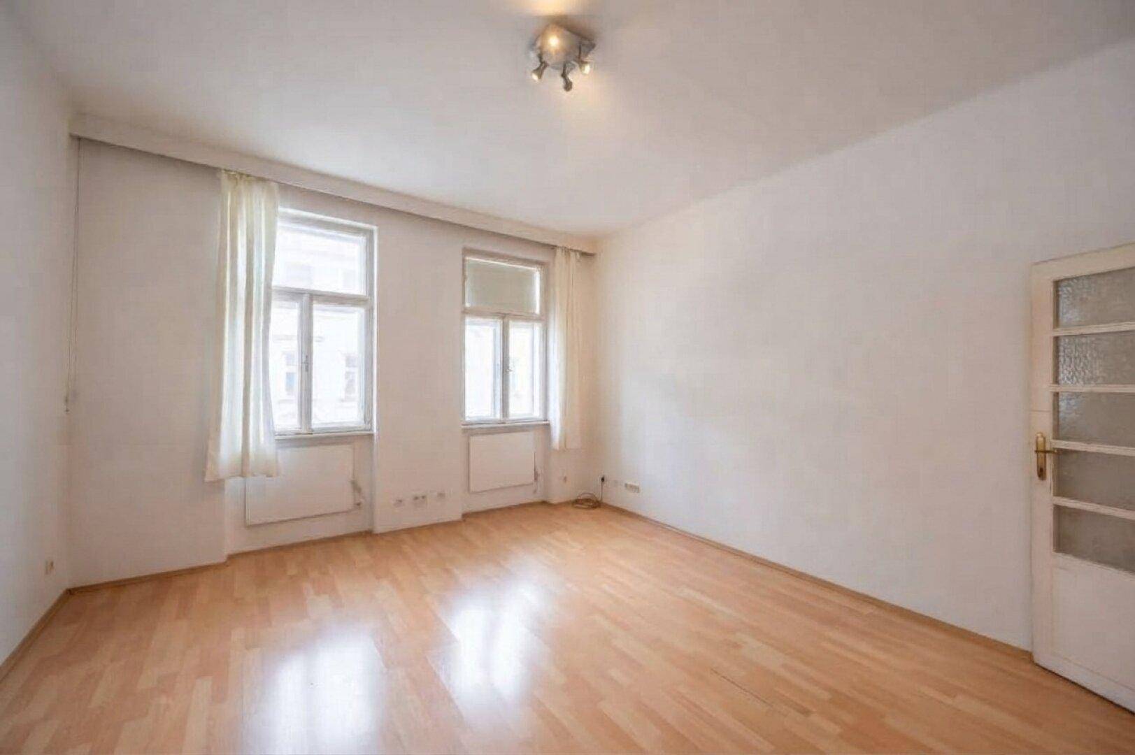 Helles Wohnzimmer mit hellem Parkettboden, weißen Wänden und zwei Fenstern.
