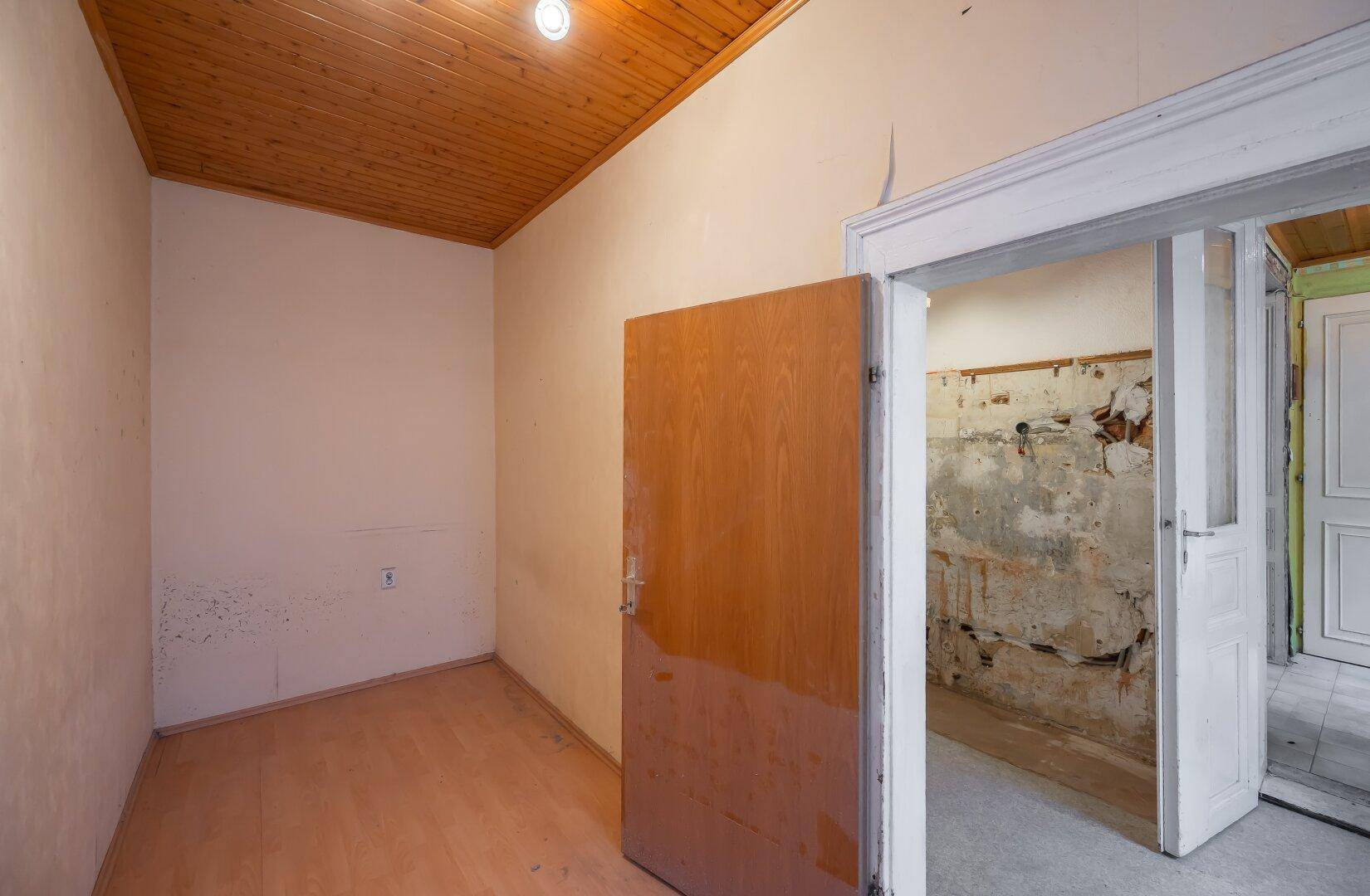 Kleiner Raum mit Holzdecke und Holztür, in renovierungsbedürftigem Zustand, mit Blick auf eine Baustelle.