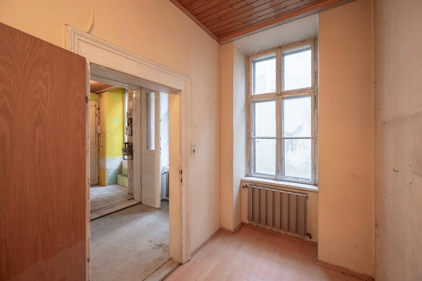 Schmaler Flur mit Fenster und Holzboden, der zu weiteren Räumen führt, in renovierungsbedürftigem Zustand.