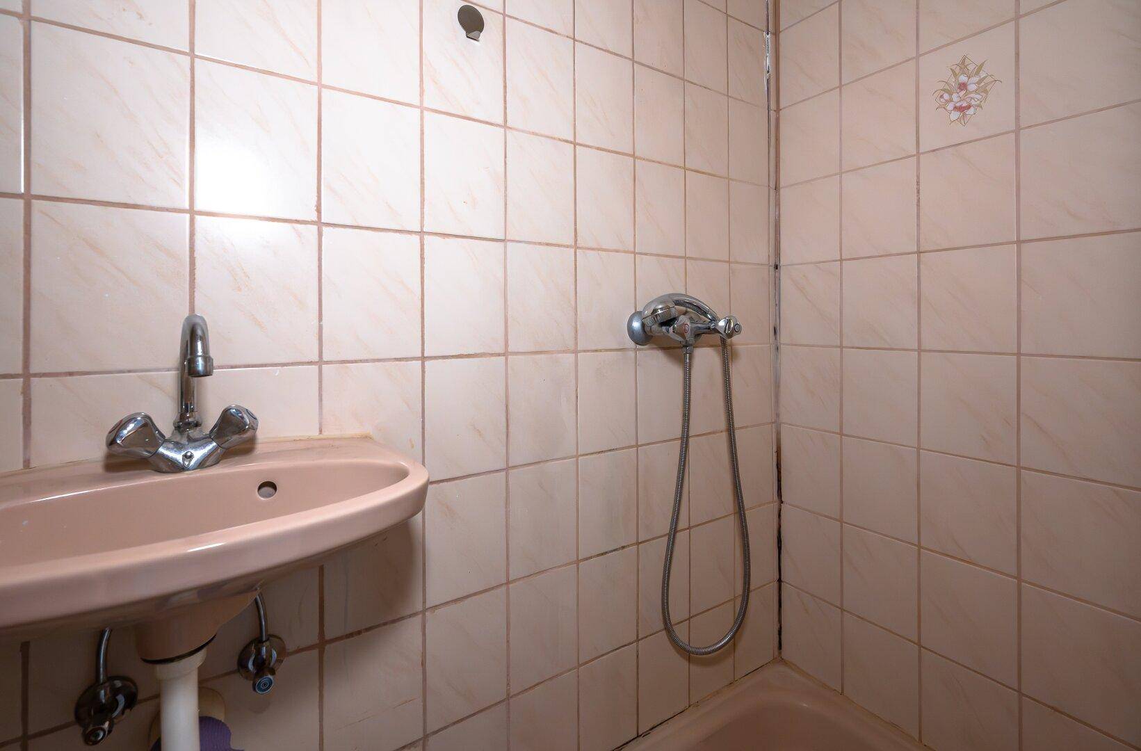 Badezimmer mit beigefarbenen Fliesen, Waschbecken und Duschanschluss, bereit zur Modernisierung.