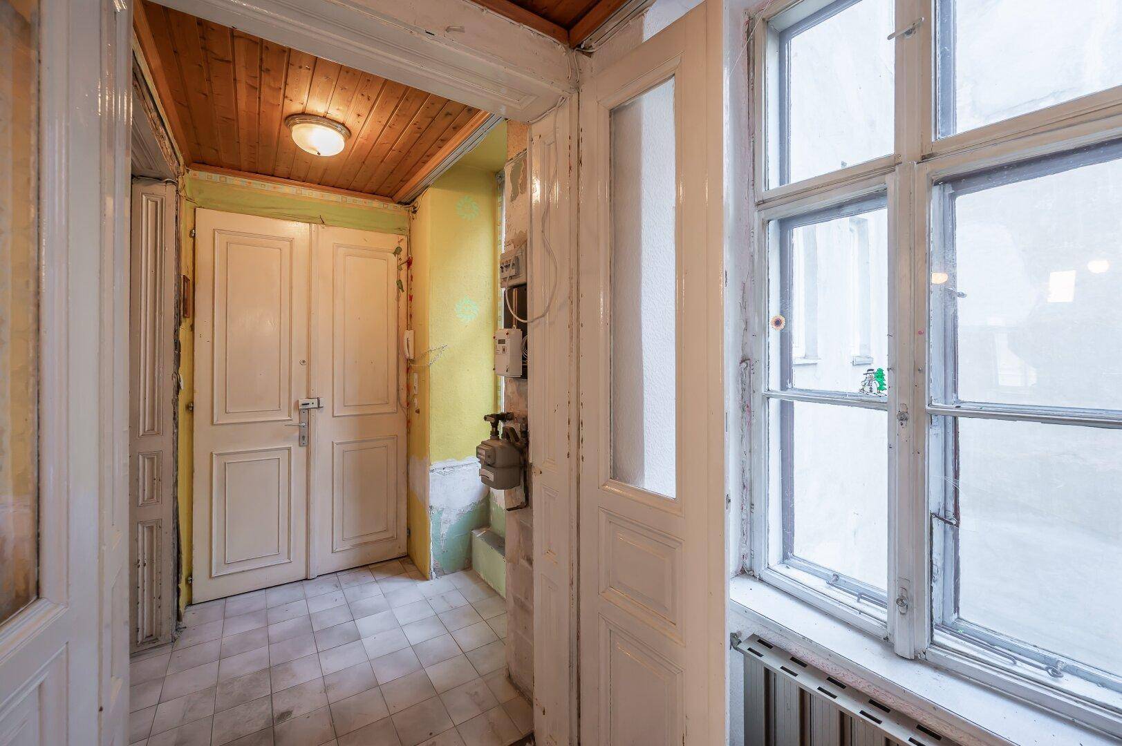 Eingangsbereich einer Wohnung mit Fliesenboden und Holzdecke, in renovierungsbedürftigem Zustand.