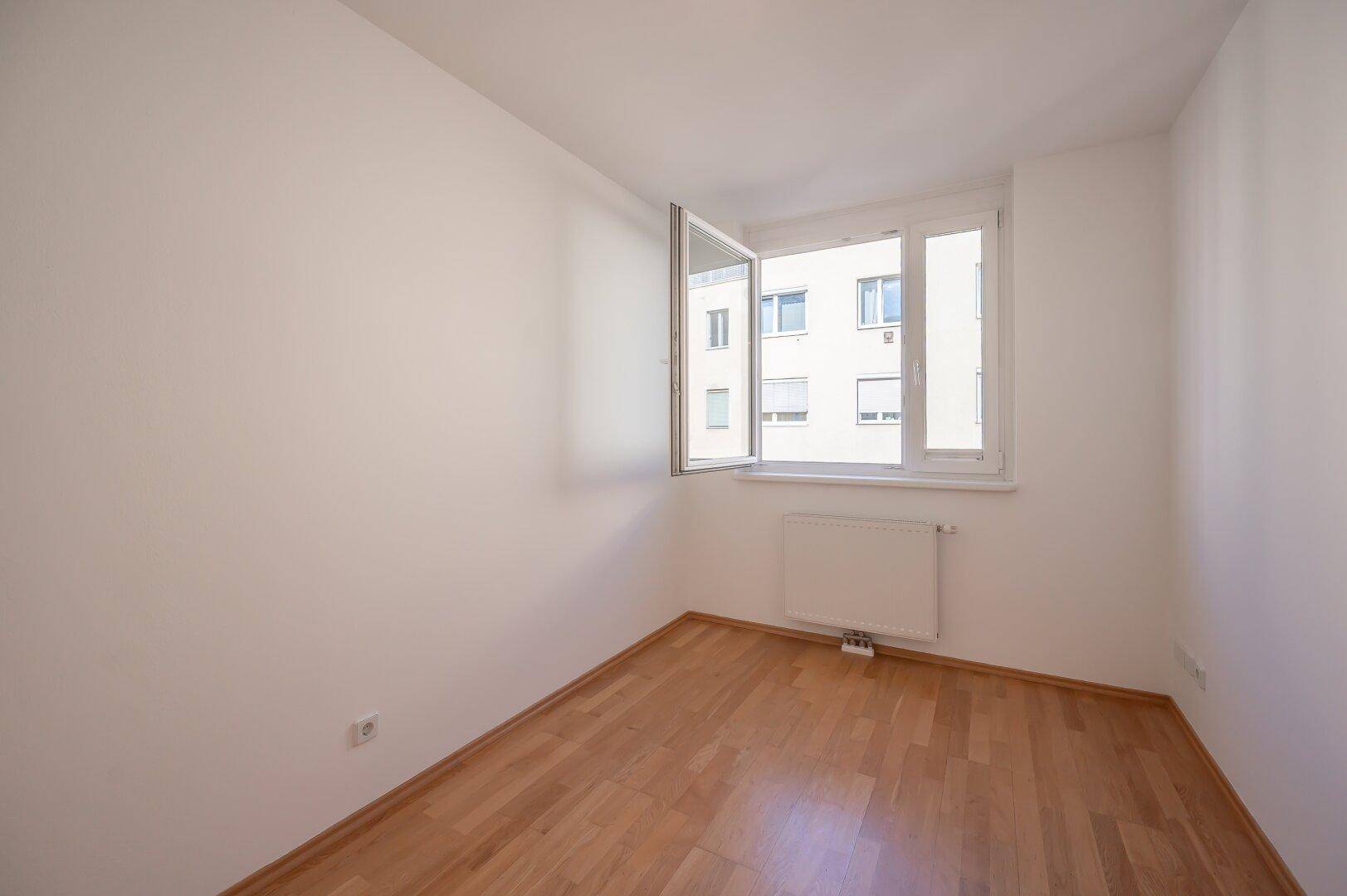 Helles Zimmer mit Parkettboden und geöffnetem Fenster, bietet viel Tageslicht und frische Luft.