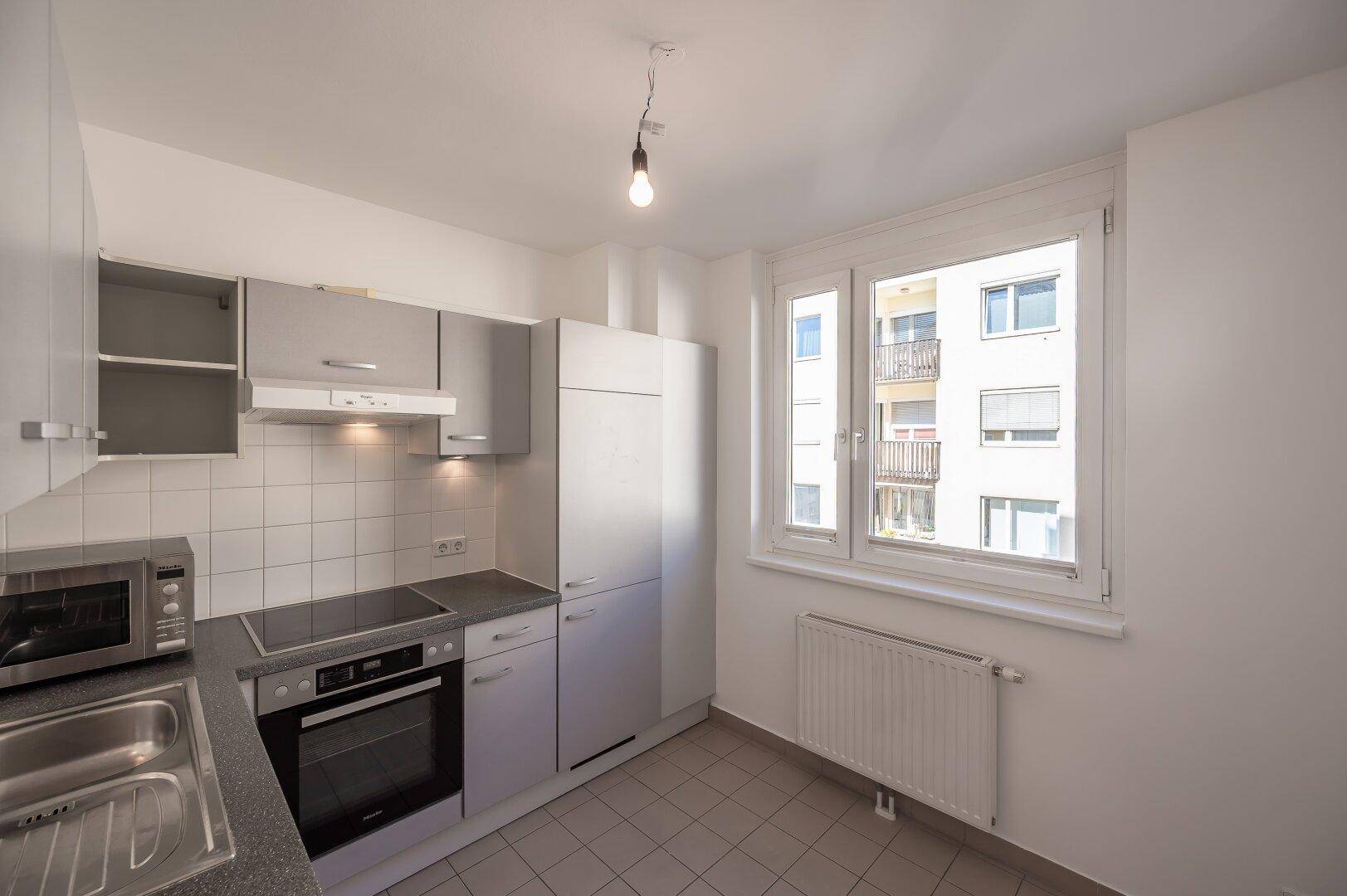 Helle Küche mit grauen Schränken, Fliesenboden und Fenster mit Blick auf die umliegenden Gebäude.