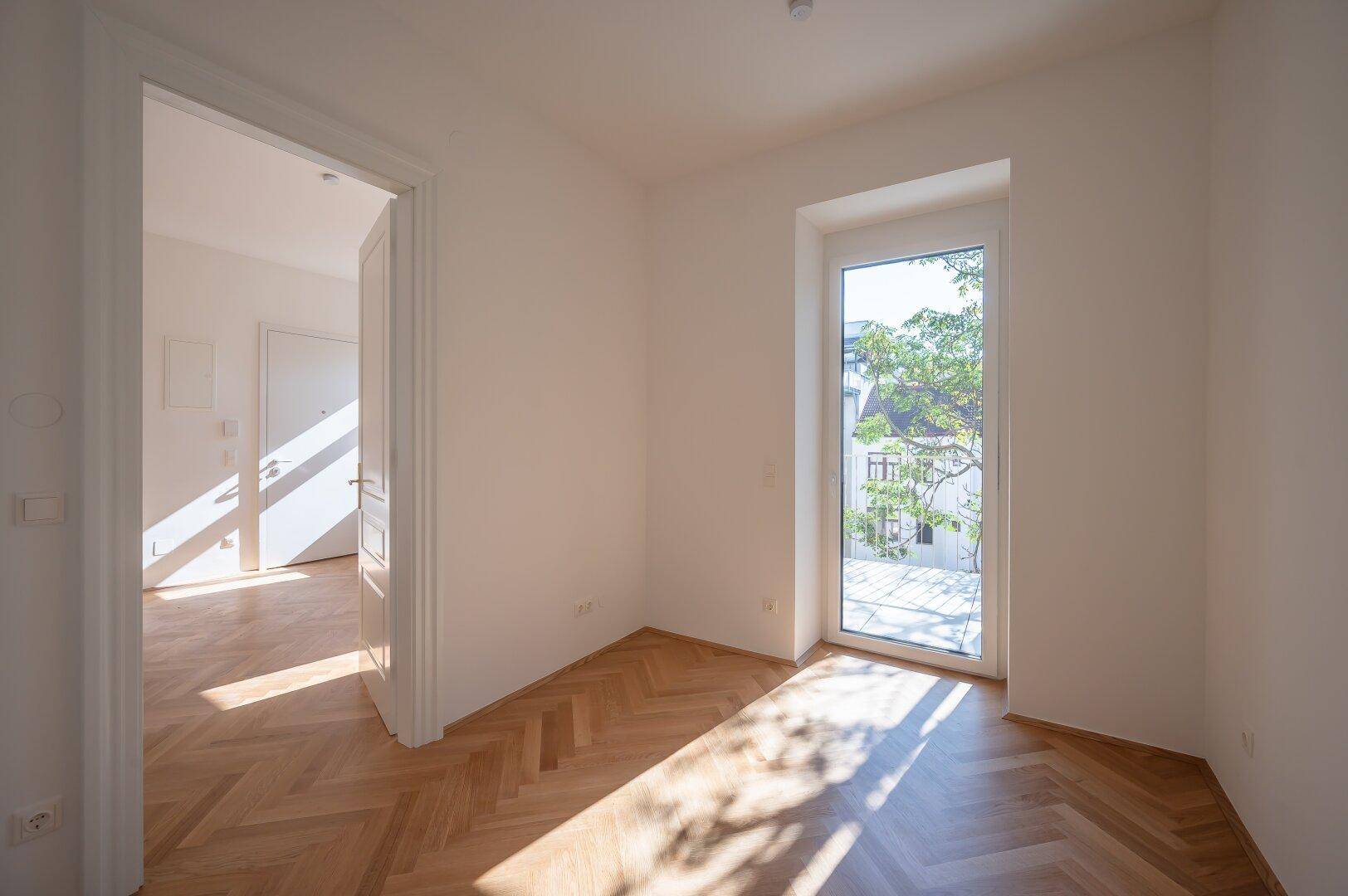 Heller Raum mit Fischgrätparkett und bodentiefem Fenster mit Blick ins Grüne.