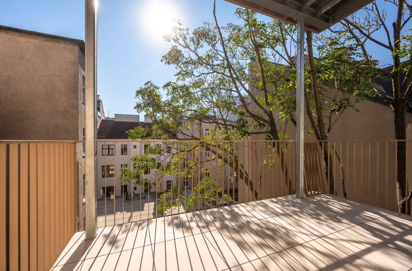Sonniger Balkon mit Fliesenboden und Blick auf den begrünten Innenhof und Bäume.