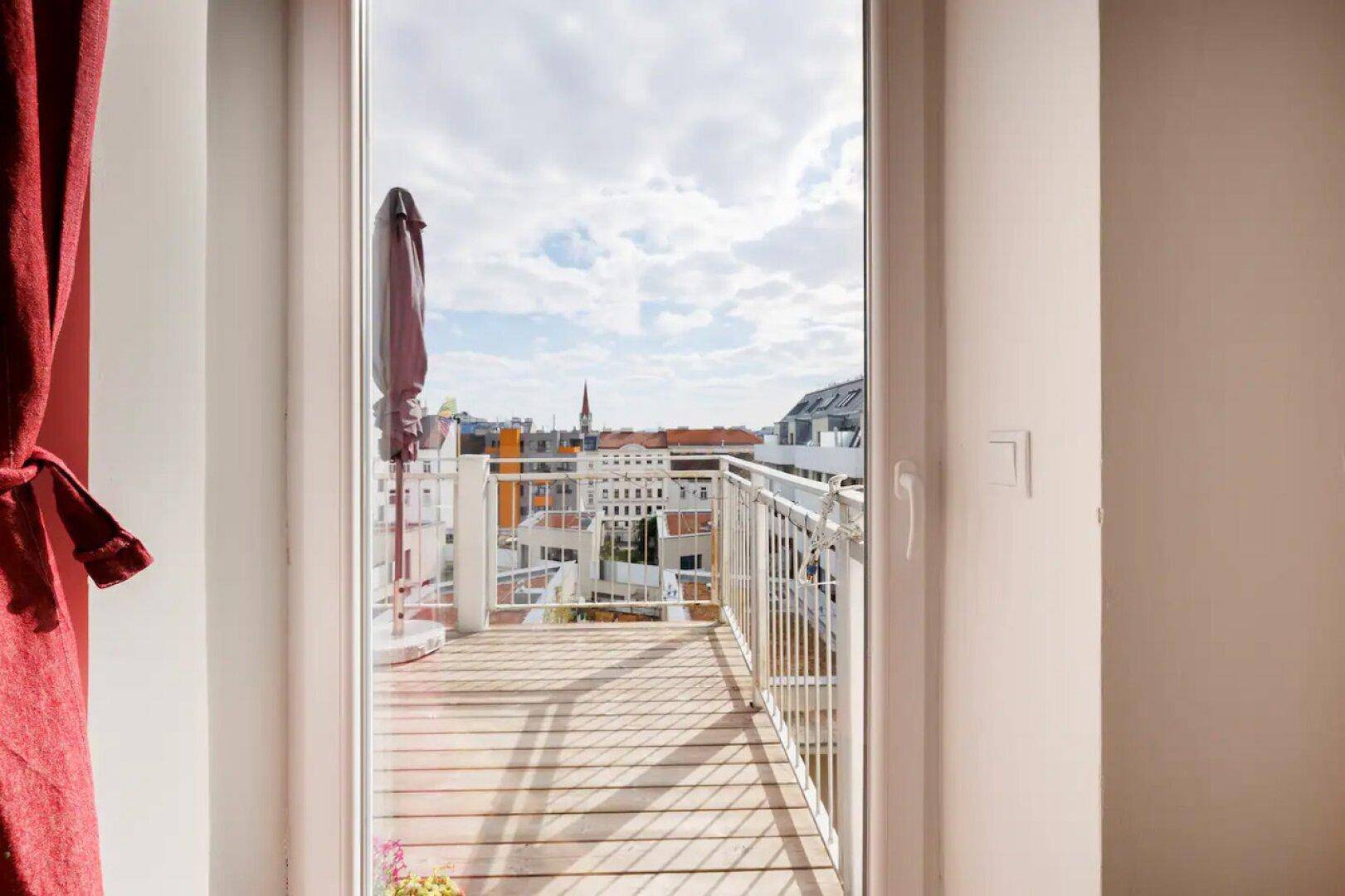 Blick vom Innenraum auf den Balkon mit Holzboden und Stadtpanorama unter bewölktem Himmel.