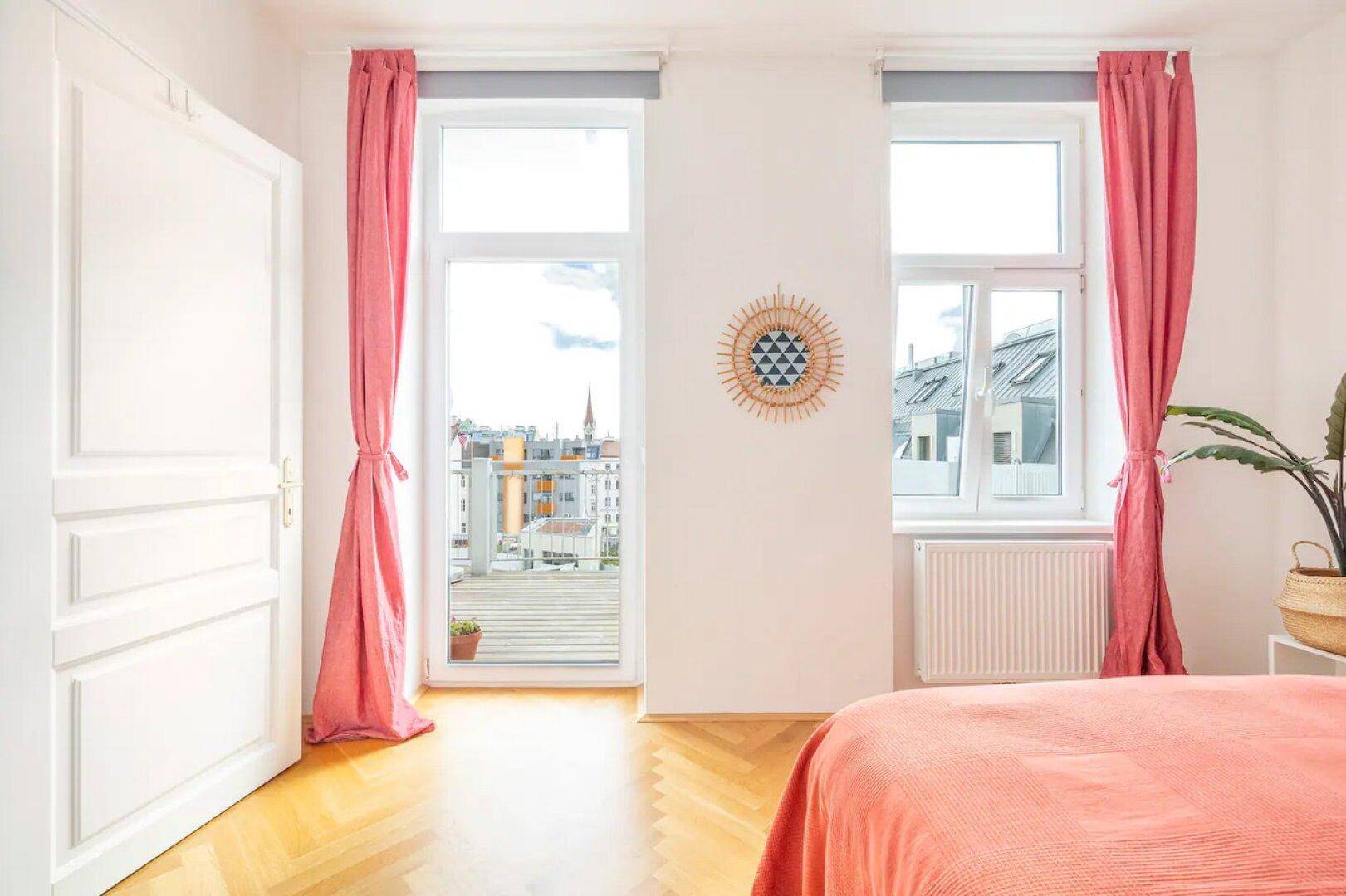 Schlafzimmer mit Parkettboden, rosa Vorhängen und direktem Zugang zu einem sonnigen Balkon.